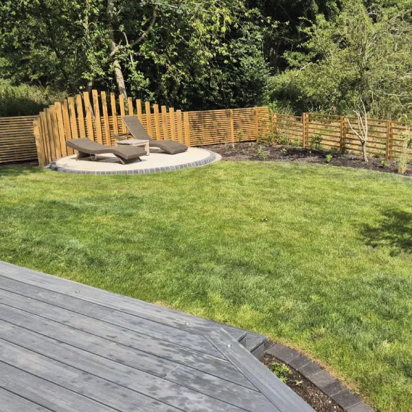 Two brown lounge chairs on a circular stone patio with wooden fencing and green lawn surrounded by trees.