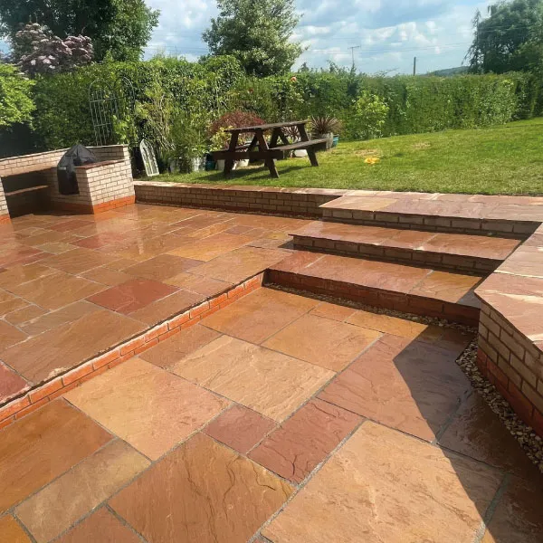 Outdoor patio with wet reddish-brown tiles, steps leading to a grass lawn with a wooden picnic table and green shrubbery.