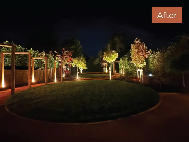 Night view of a landscaped garden with a circular grass lawn, pathway, and illuminated trees and wooden pergolas.