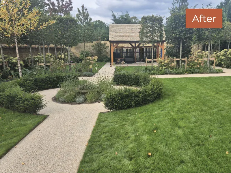 Landscaped garden with gravel paths, lush grass, trimmed bushes, young trees, and a wooden gazebo against a stone wall.