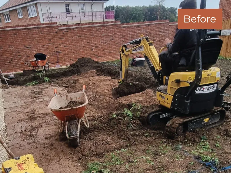 Person operating mini excavator digging soil in a backyard with wheelbarrows and a brick wall.