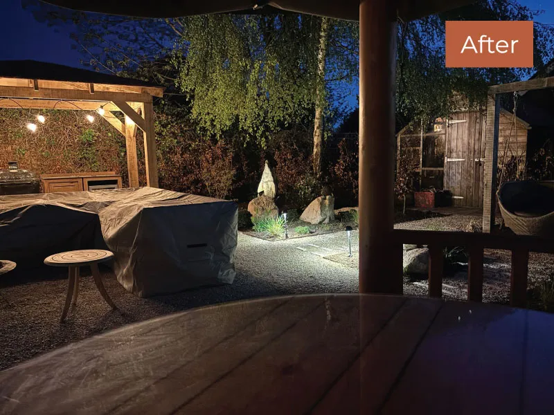 Night view of a backyard with covered furniture, illuminated garden statue, and a wooden shed under a pergola.