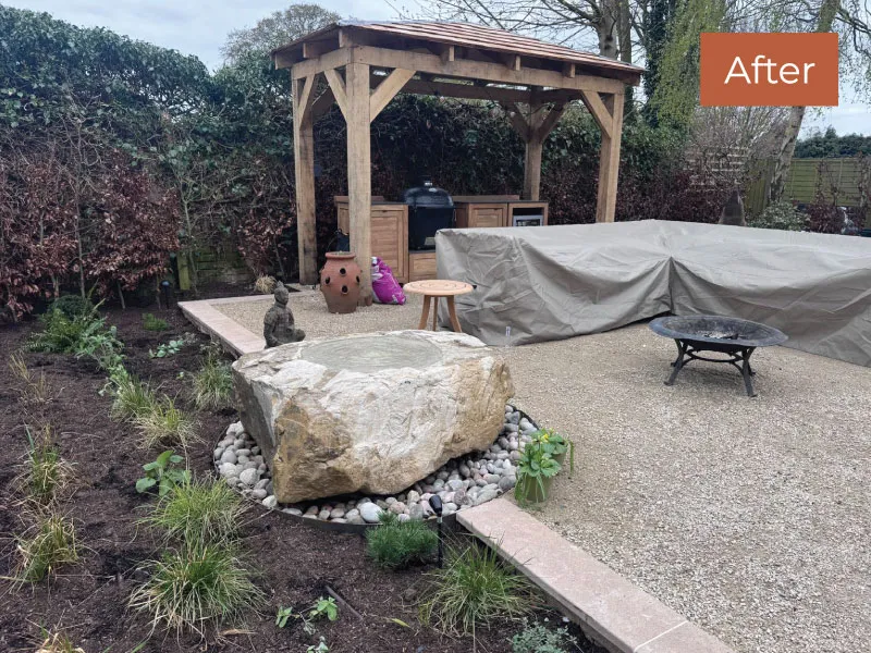 Backyard patio with a wooden pergola, covered furniture, fire pit, and a large stone water feature surrounded by plants.