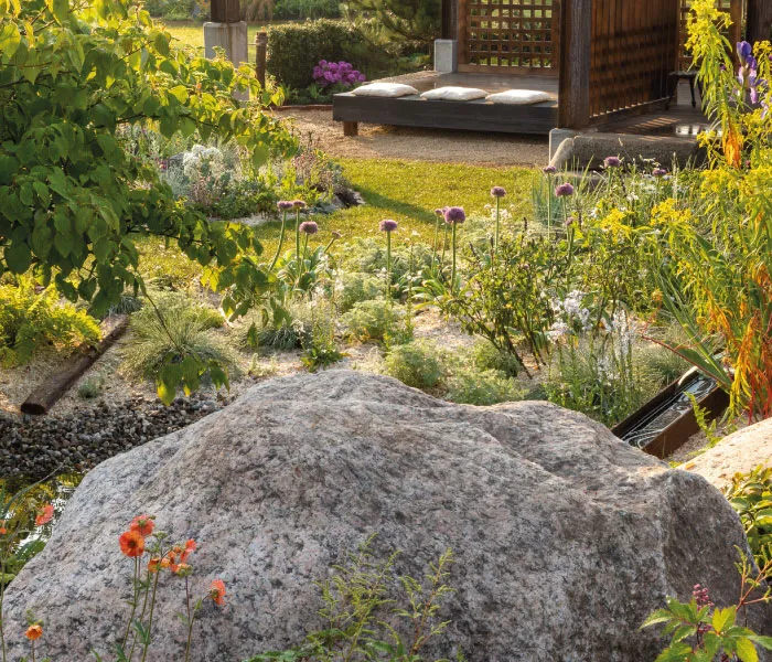 Sunlit garden with a large rock, blooming flowers, green foliage, and a shaded wooden bench with cushions.