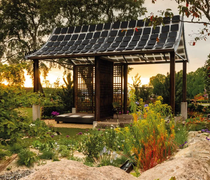 Open wooden garden pavilion with solar panels on roof surrounded by colorful flowers and greenery at sunset.
