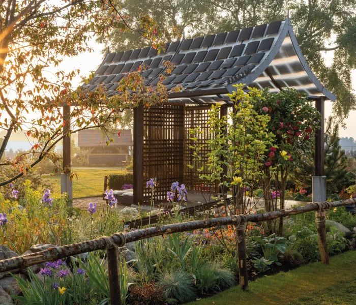 Wooden garden pavilion with a curved roof surrounded by flowering plants and a rustic wooden fence.