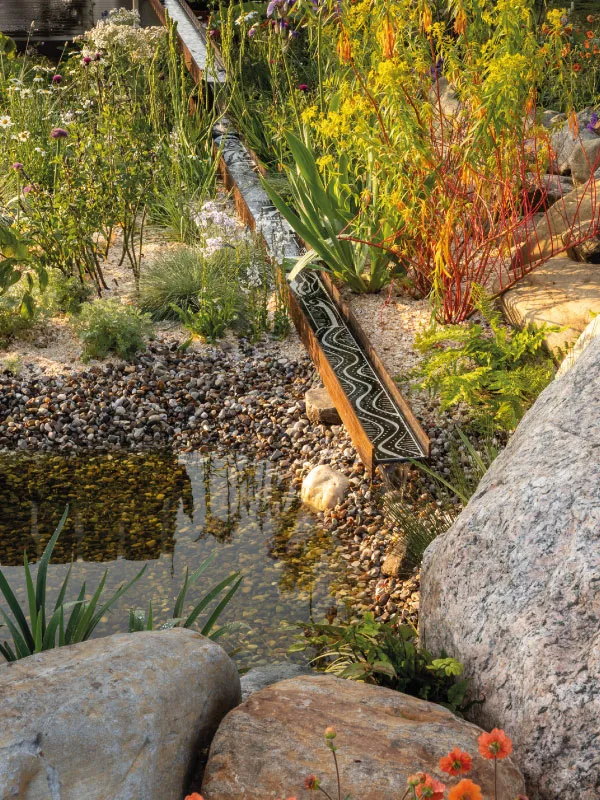 Rocky garden with a narrow water channel running through colorful plants beside a small pond.