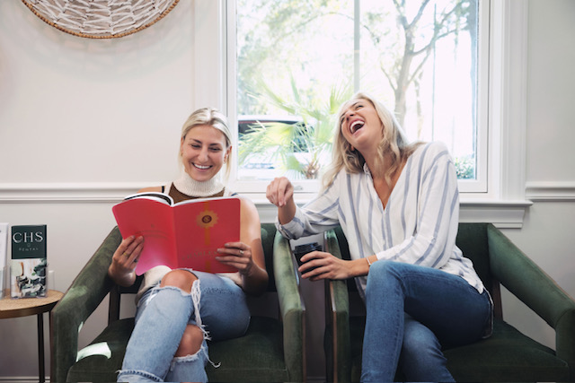 Two women sitting on green chairs by a window, one reading a red book and the other laughing holding a cup.