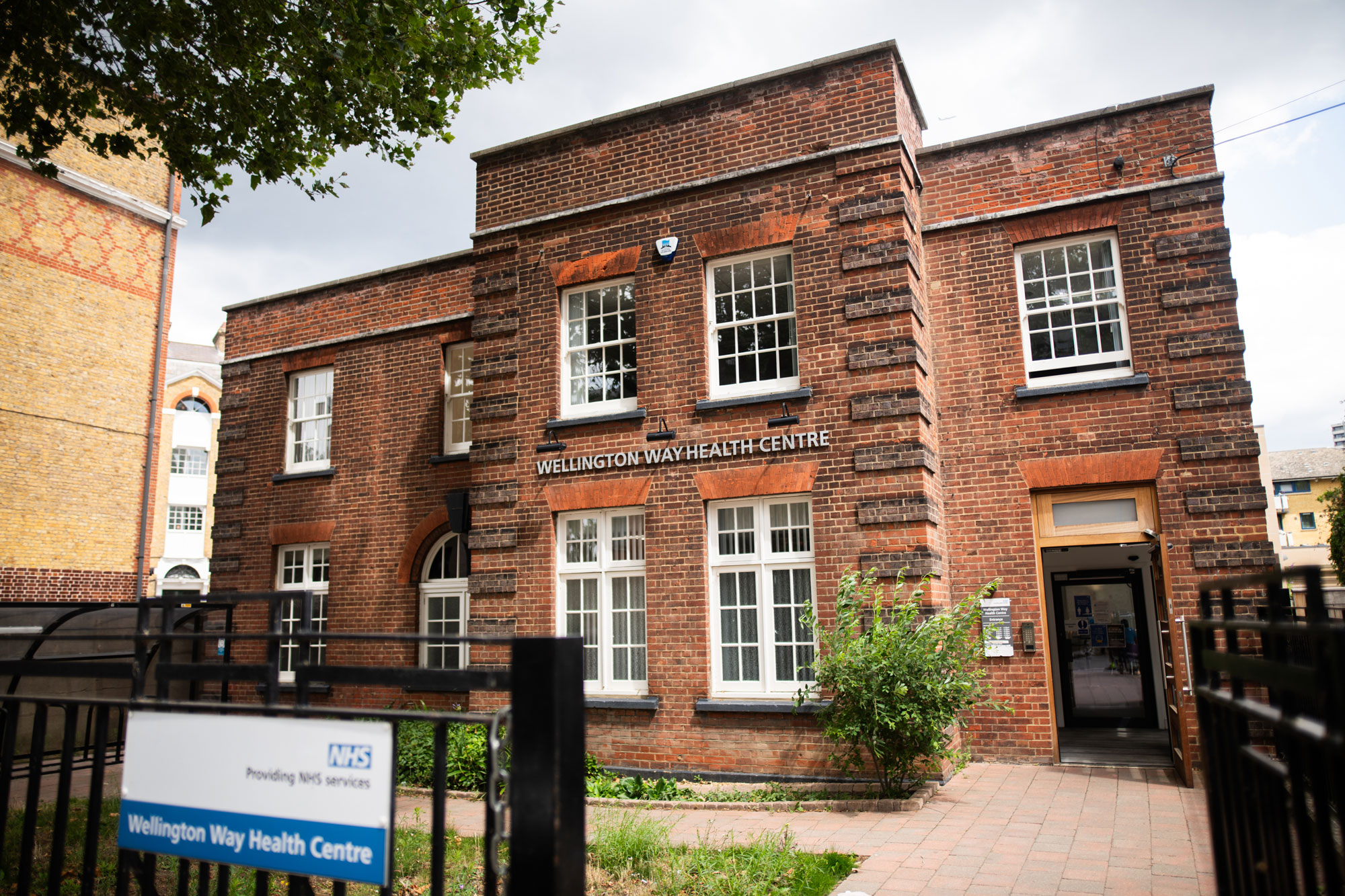 Red brick Wellington Way Health Centre building with open entrance door and fenced pathway.