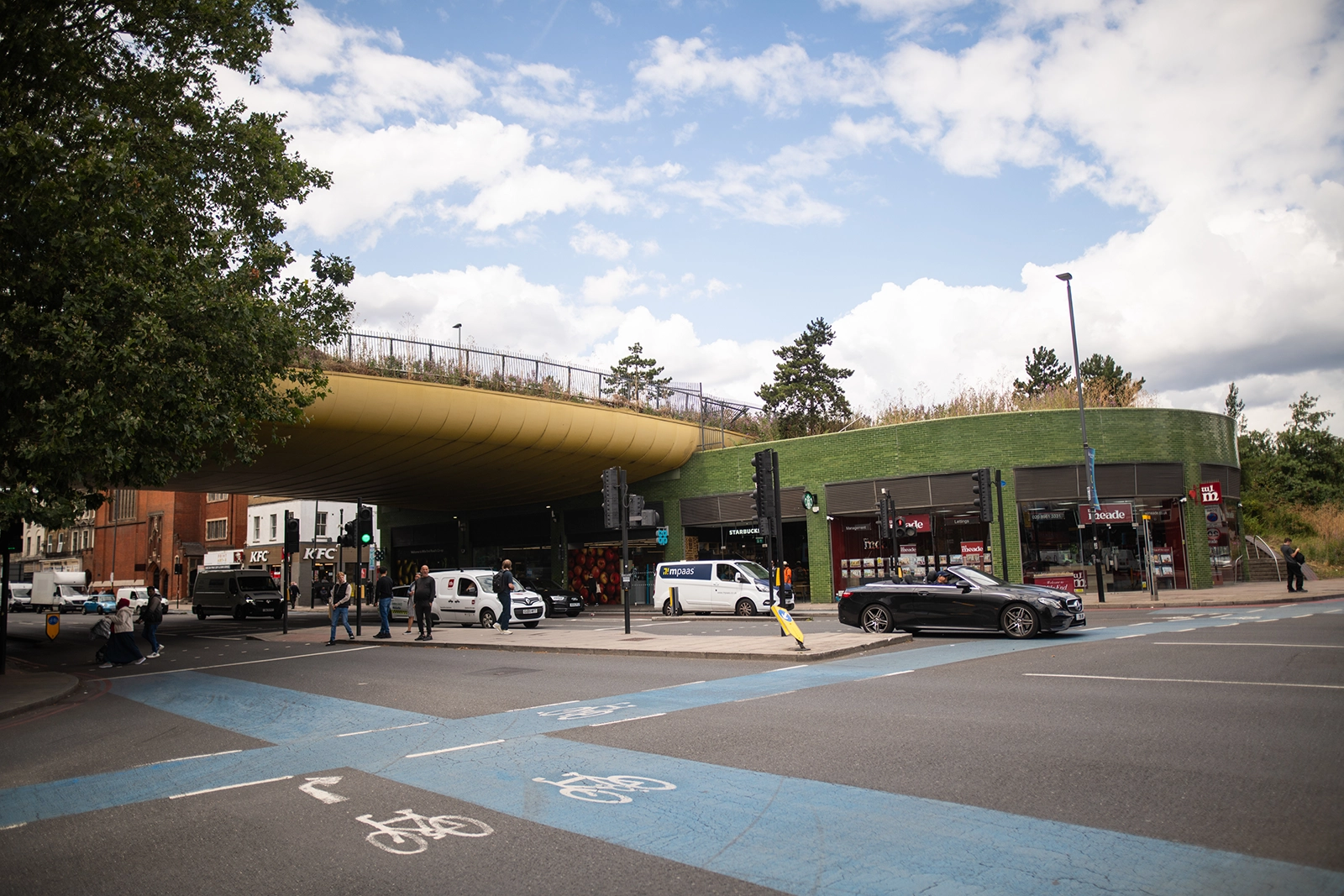 Street intersection with bike lanes, vehicles, pedestrians, green-roofed building, and partly cloudy sky.