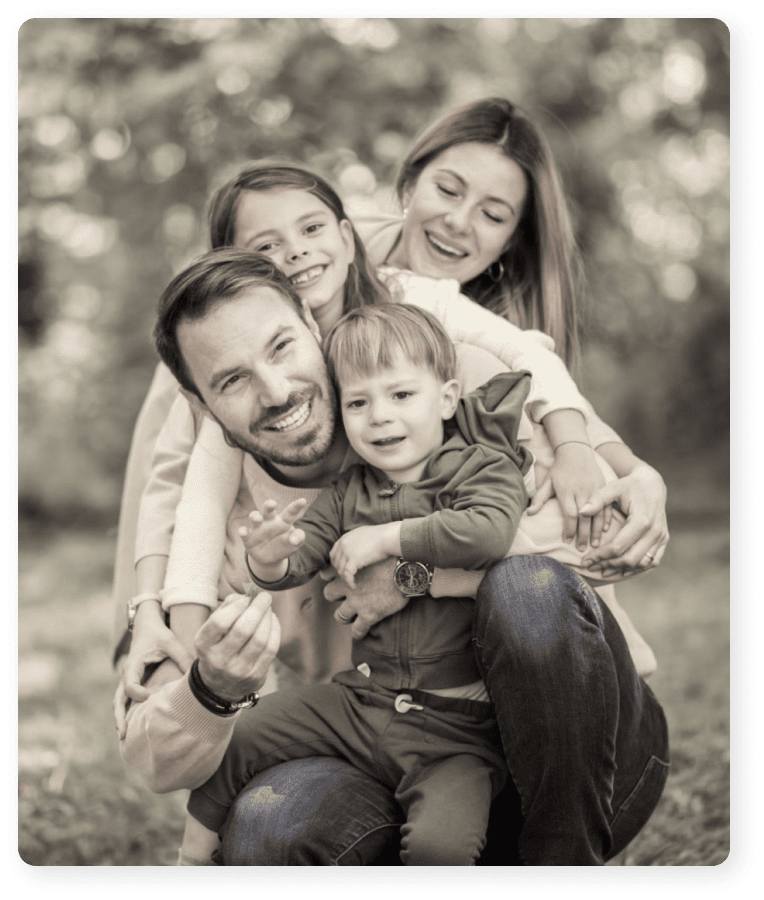 Smiling family of four hugging outdoors with father crouching and children and mother leaning on him.