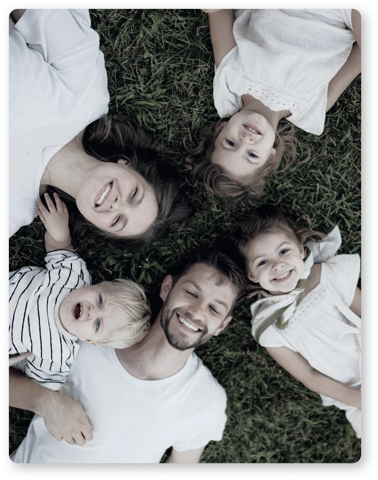 Smiling family of five lying on grass in a circle, with adults and children looking up.