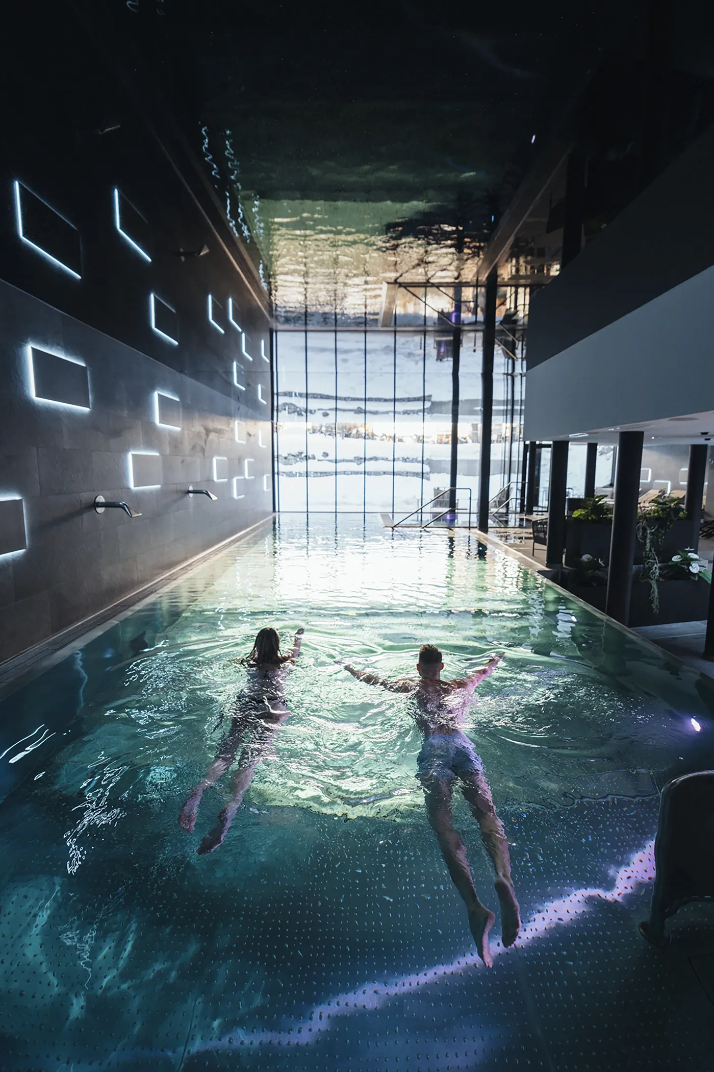 Paar schwimmt im modernen Hallenbad des Alpen Resort & Spa Zermatt, Wellness-Bereich mit beleuchteter Wand und Blick auf den Schnee durch grosse Fensterfronten.