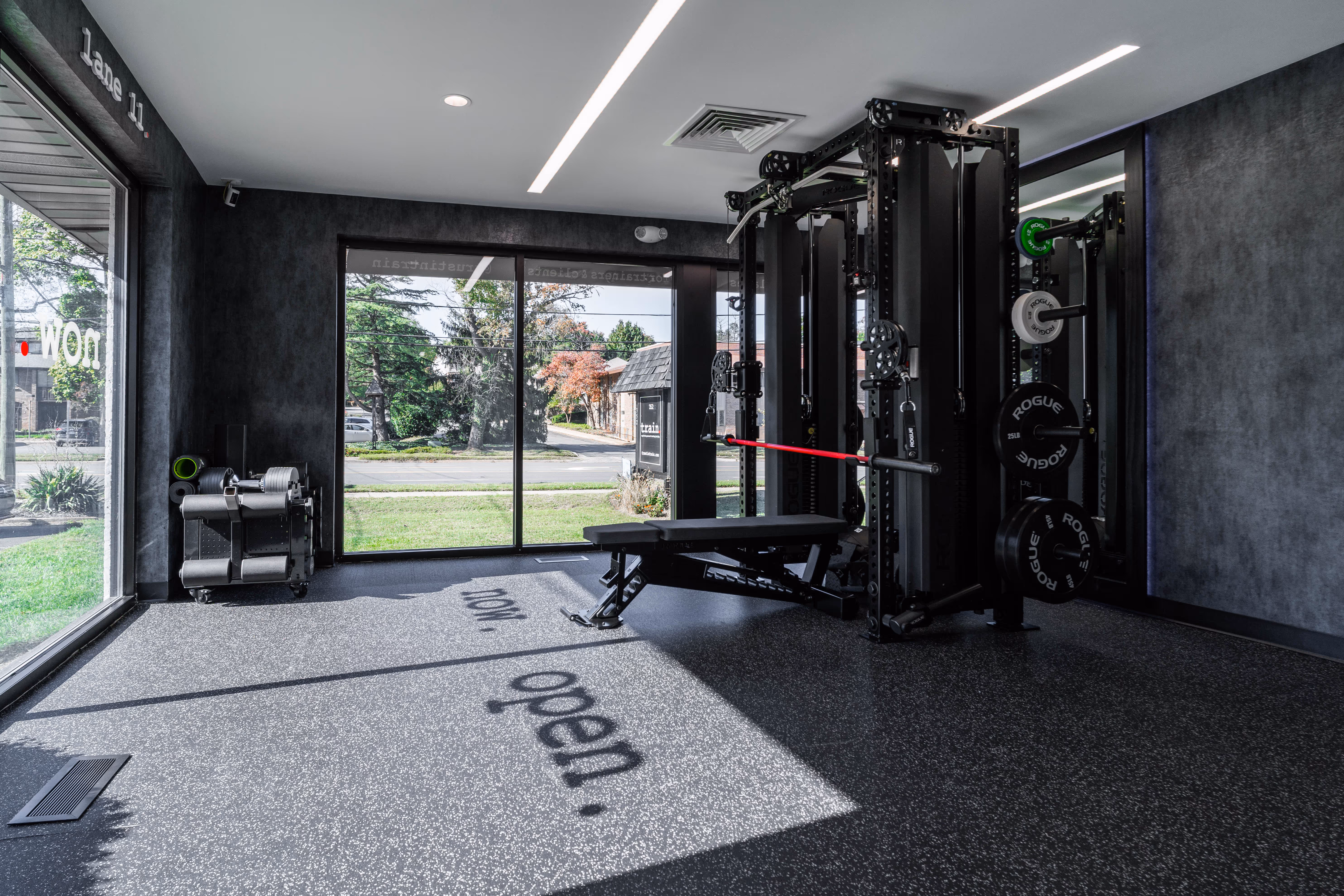Modern gym room with exercise bench, weight rack, and large windows showing green outdoor area.