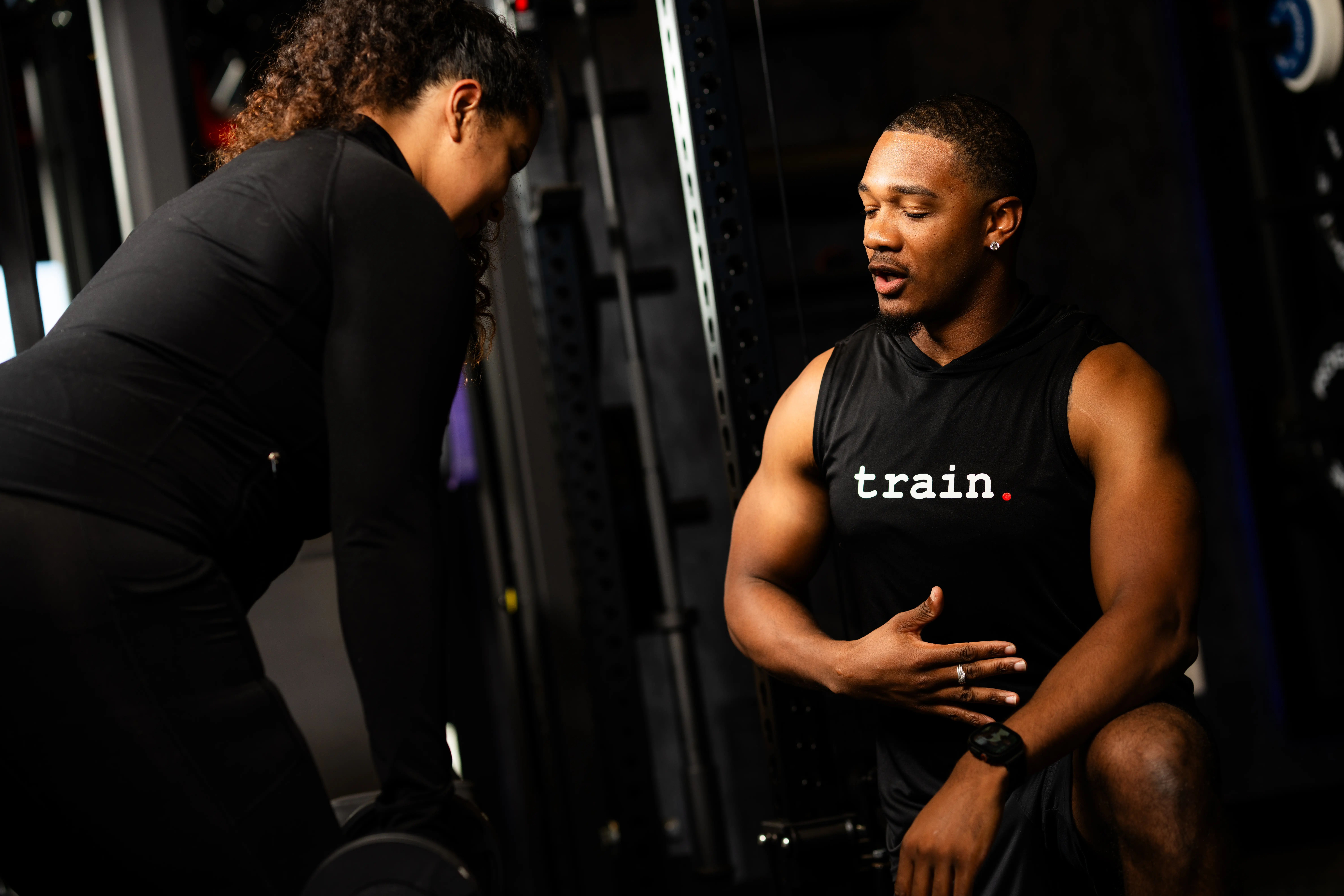Muscular man in a sleeveless shirt coaching a woman lifting a dumbbell in a gym.
