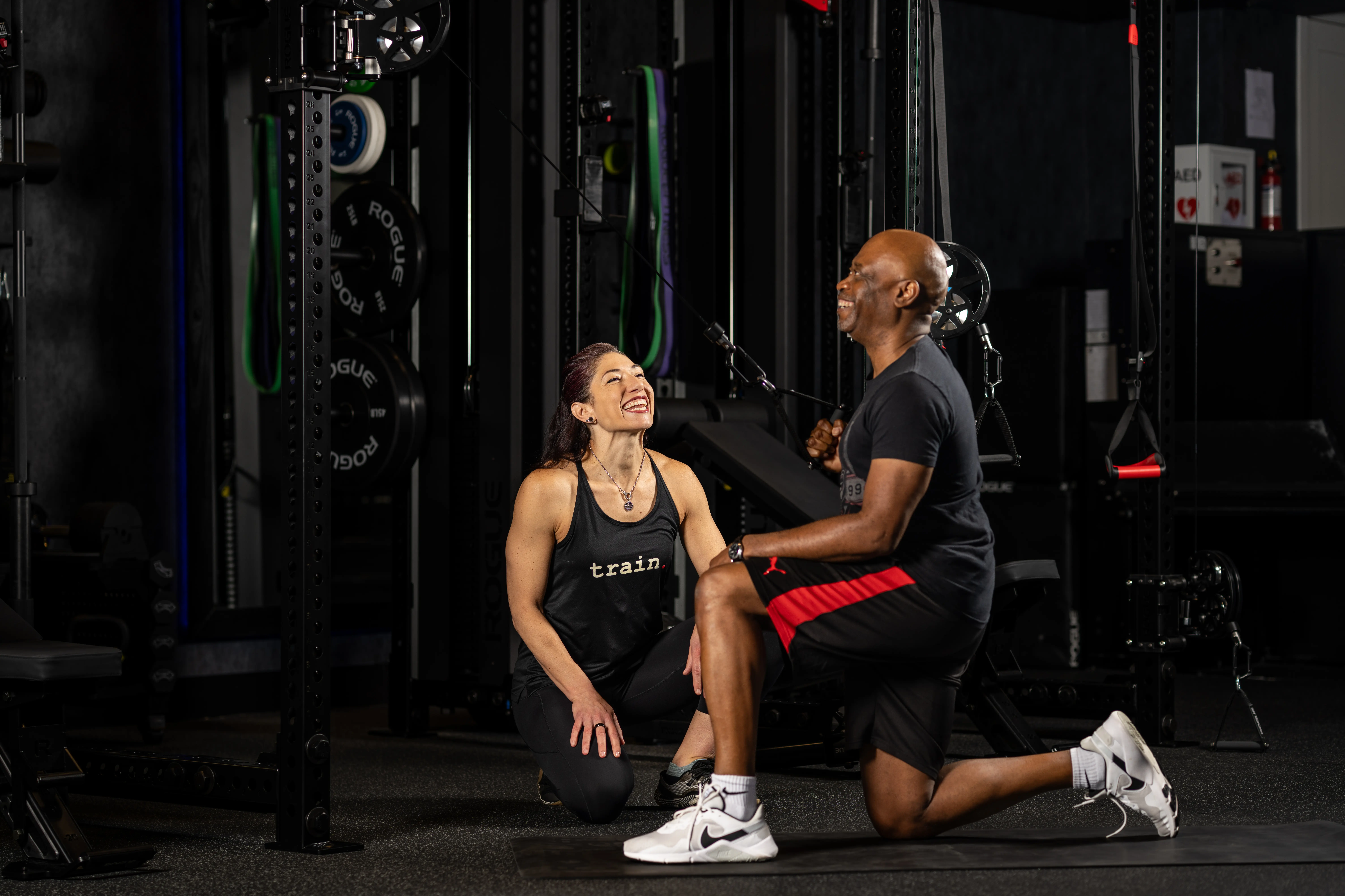 Man doing cable workout while trainer kneels nearby, smiling, in a gym setting with weights and equipment.