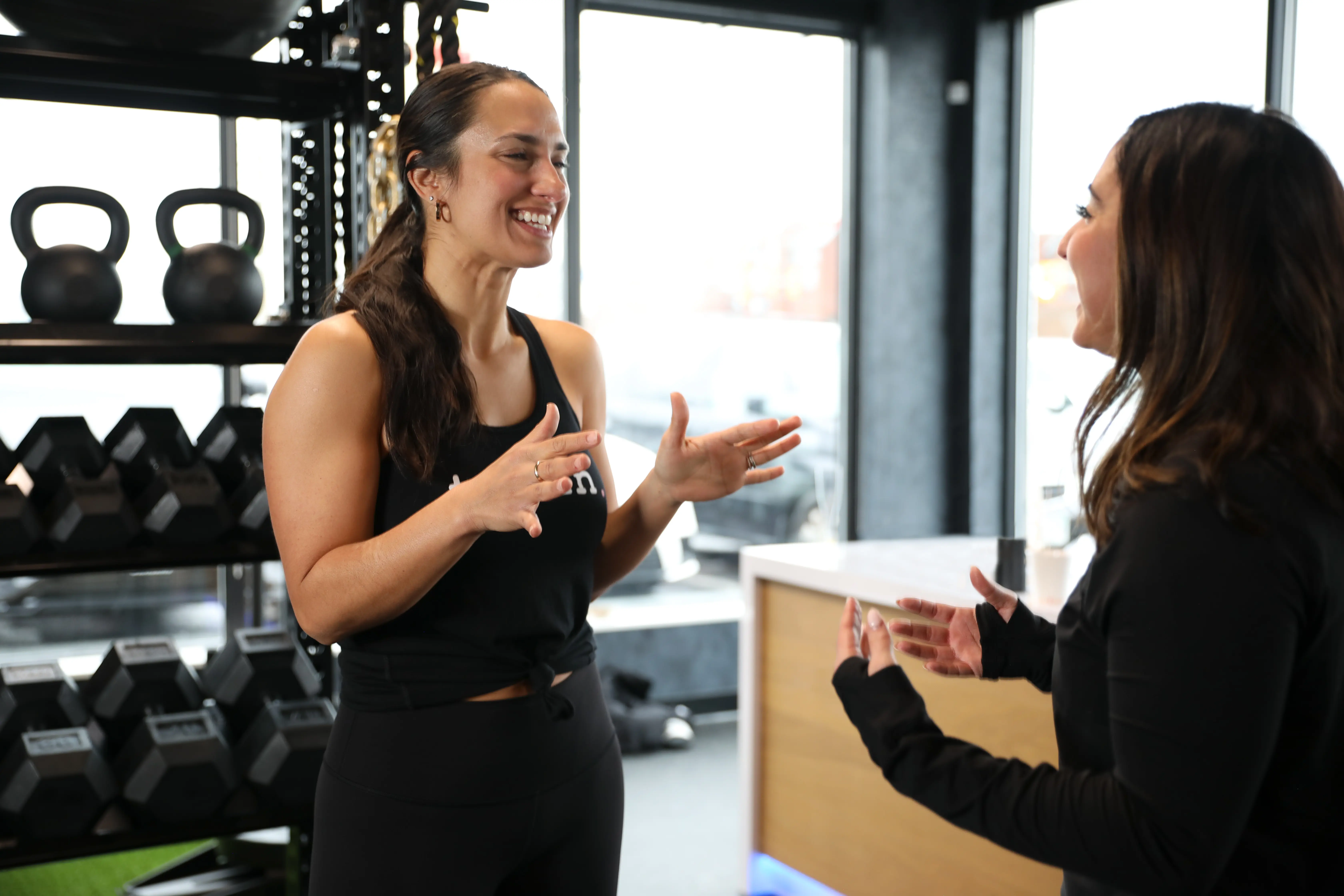 Two women smiling and talking in a gym near dumbbells and kettlebells on shelves.