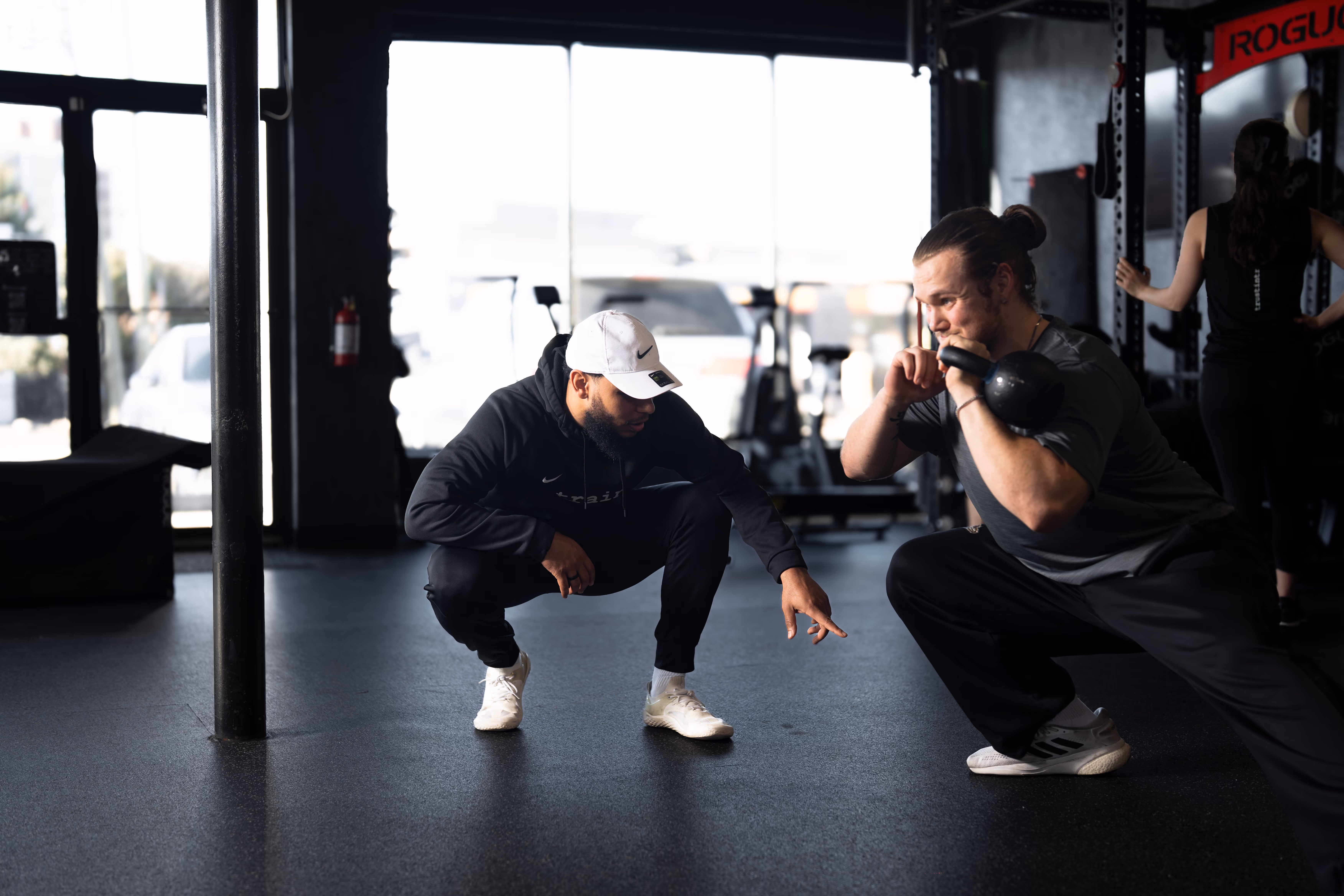 Trainer crouching and coaching a man lifting a kettlebell in a gym workout session.