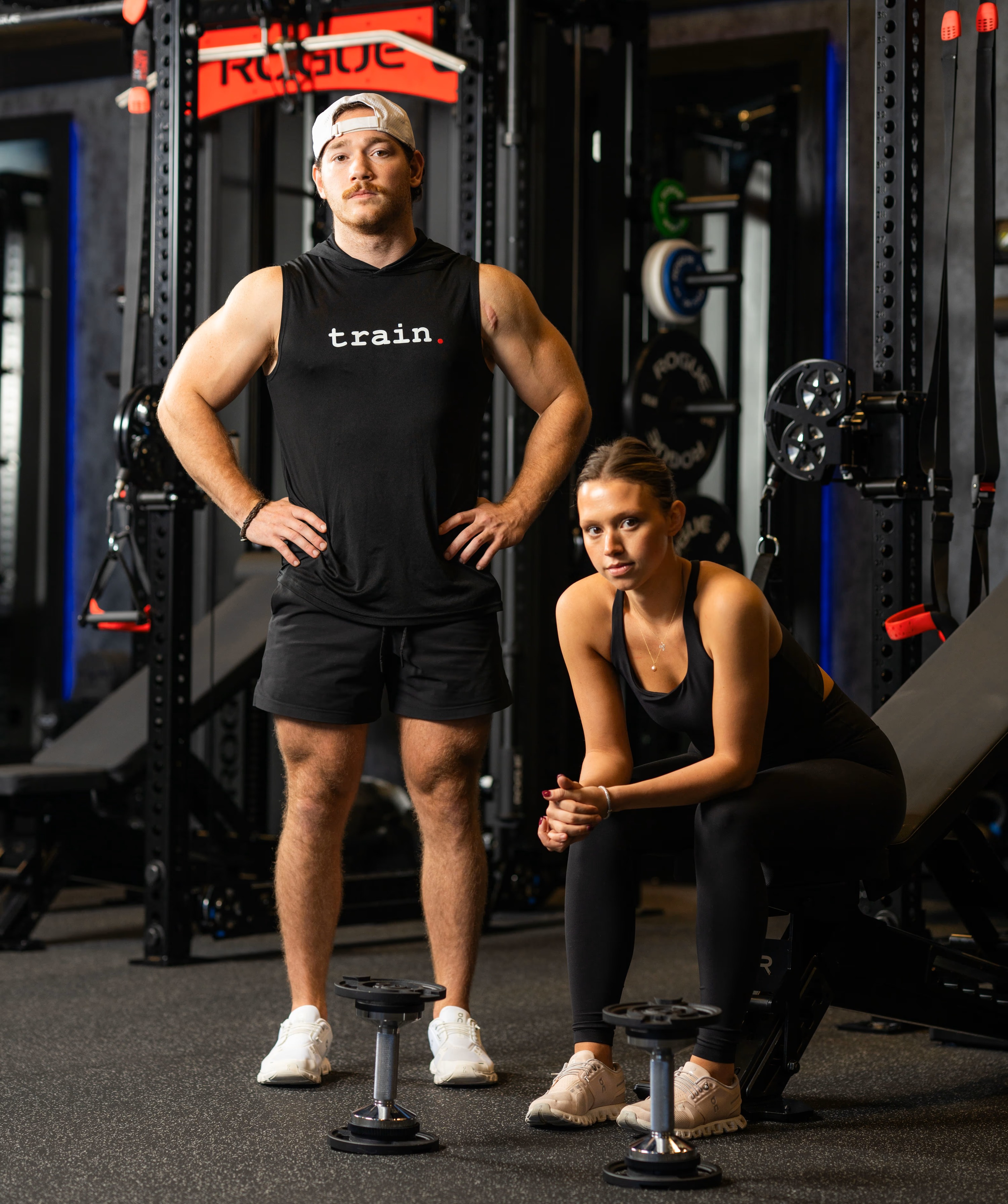 Man standing with hands on hips and woman sitting on bench in gym with dumbbells on floor.