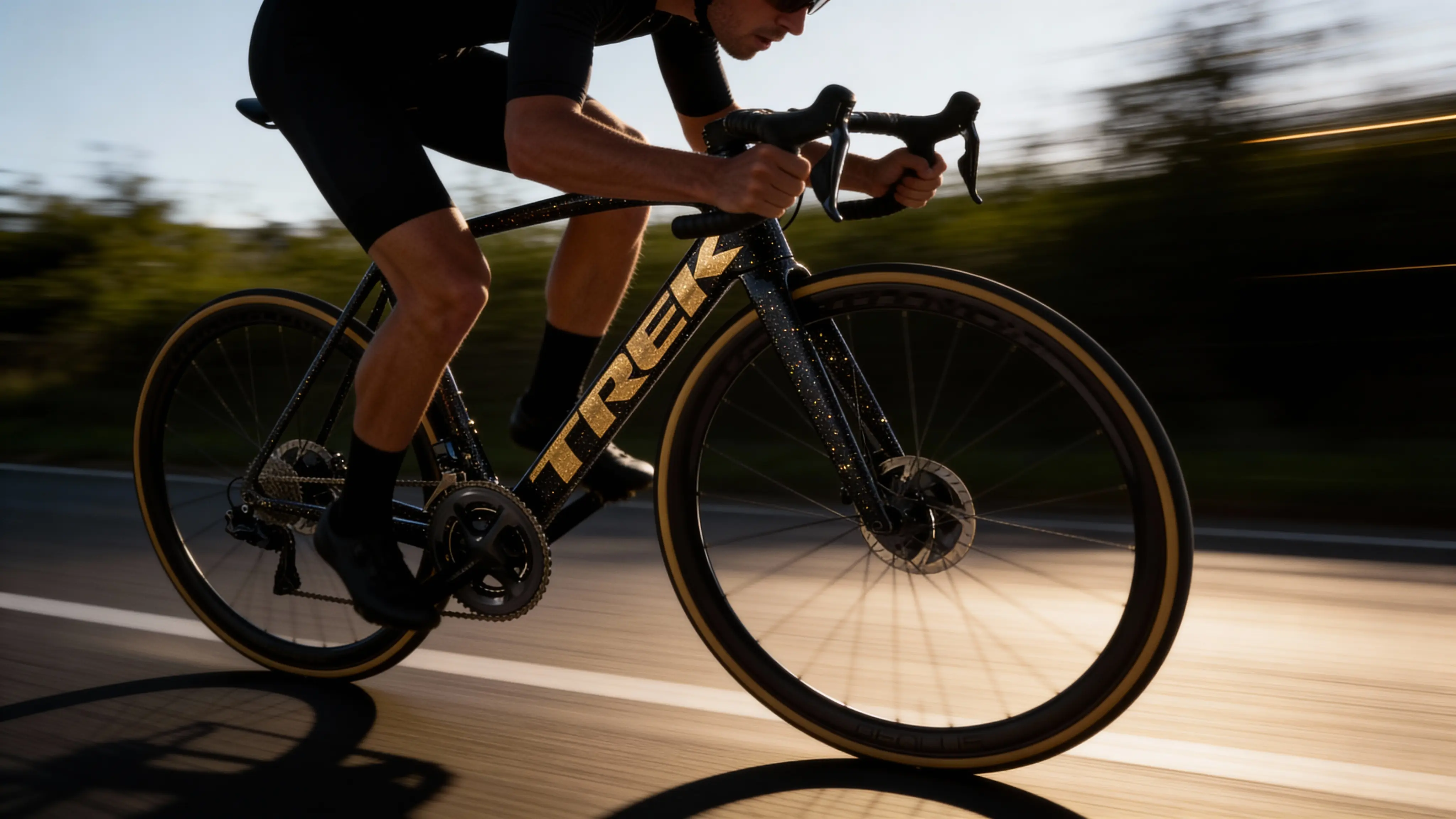 Cyclist riding a black and gold Trek road bike on a paved road at sunset.