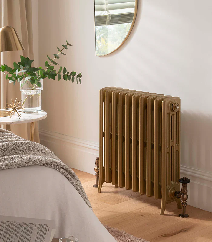Bronze vintage-style radiator on wood floor next to bed with beige blanket and a small white round table with a glass vase and green leaves.