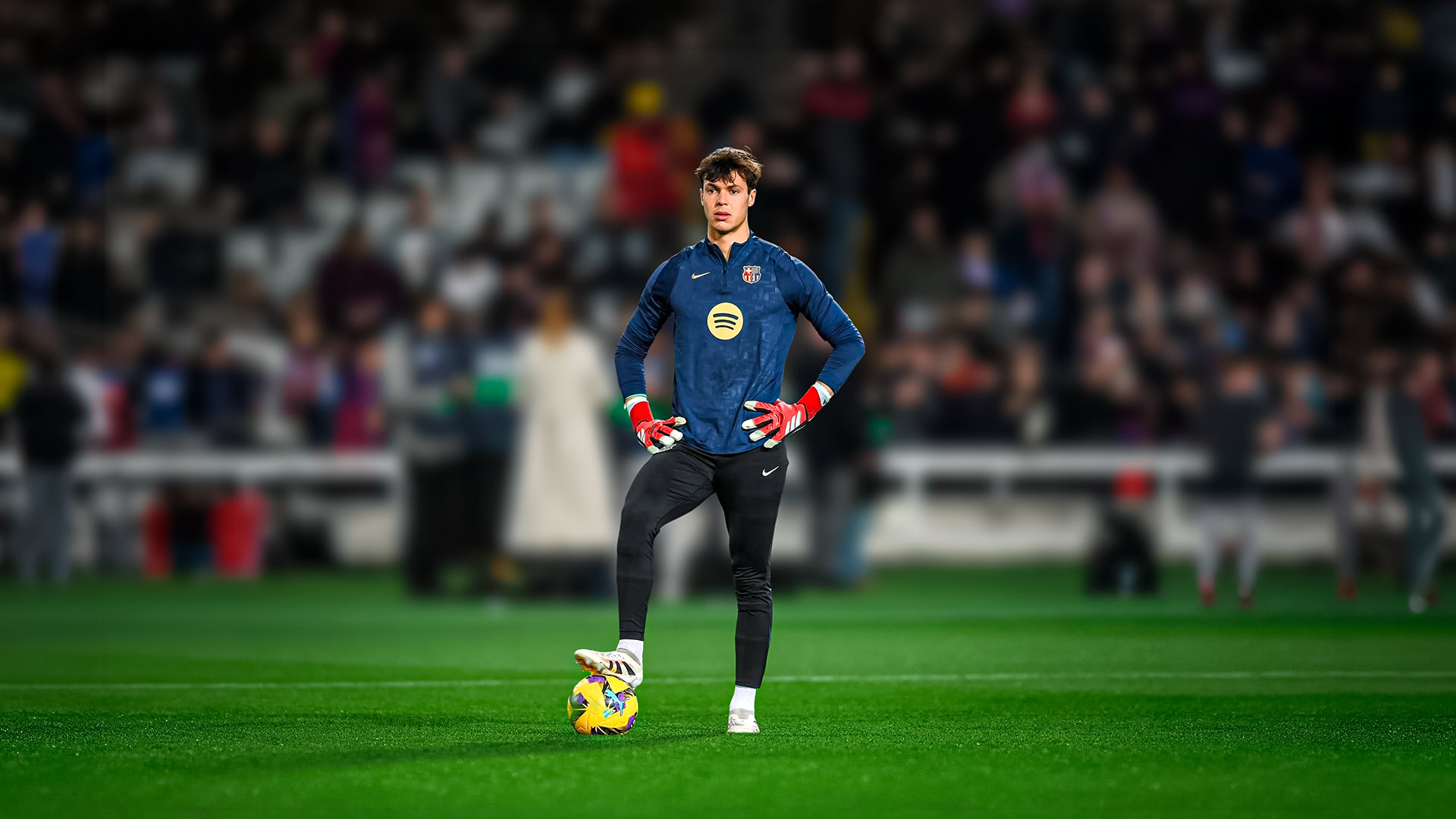 Soccer goalkeeper in blue training top stands on grass with one foot on a yellow ball in a stadium.