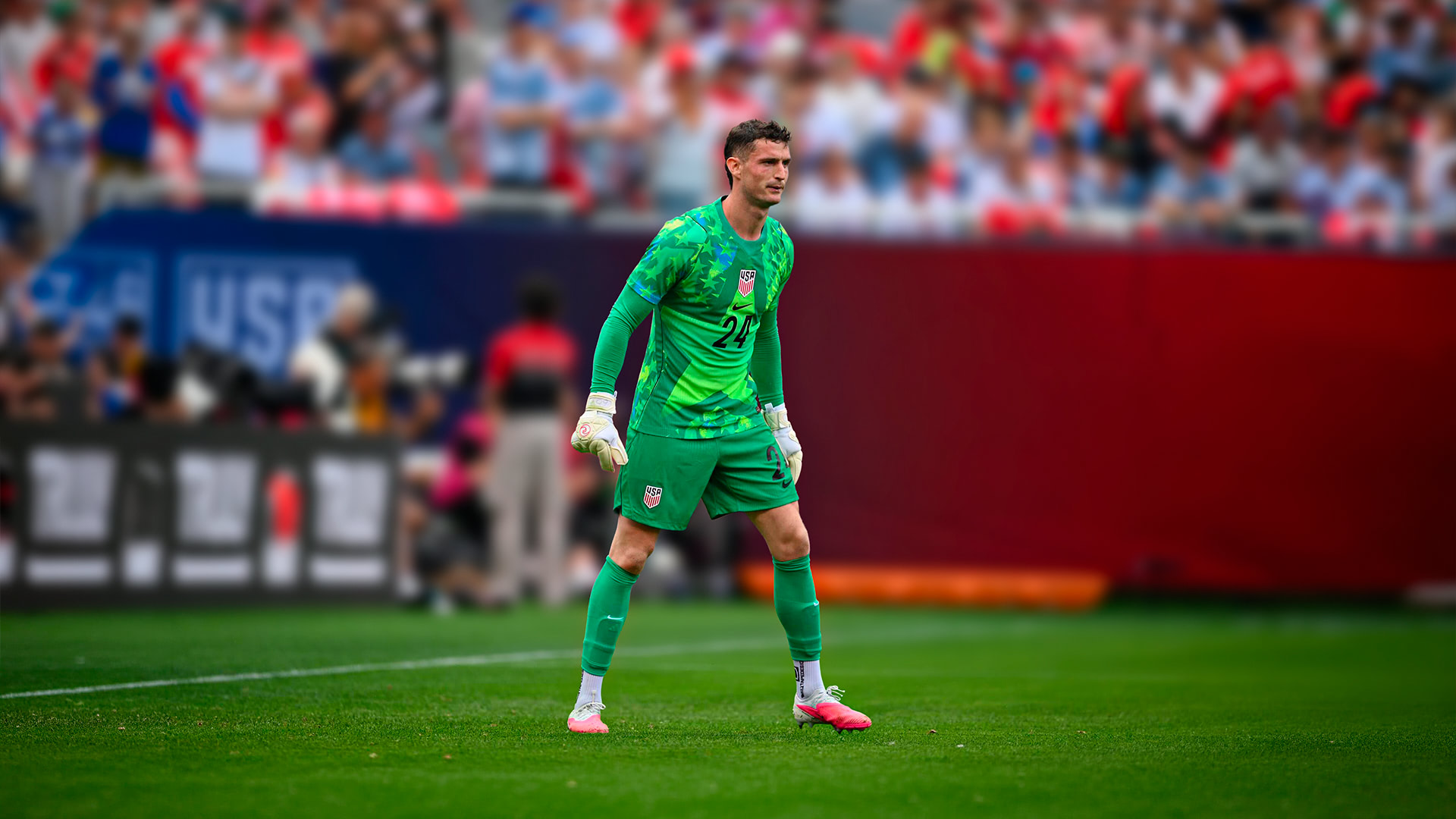 Soccer goalkeeper in green USA uniform on field during a match with blurred crowd behind.