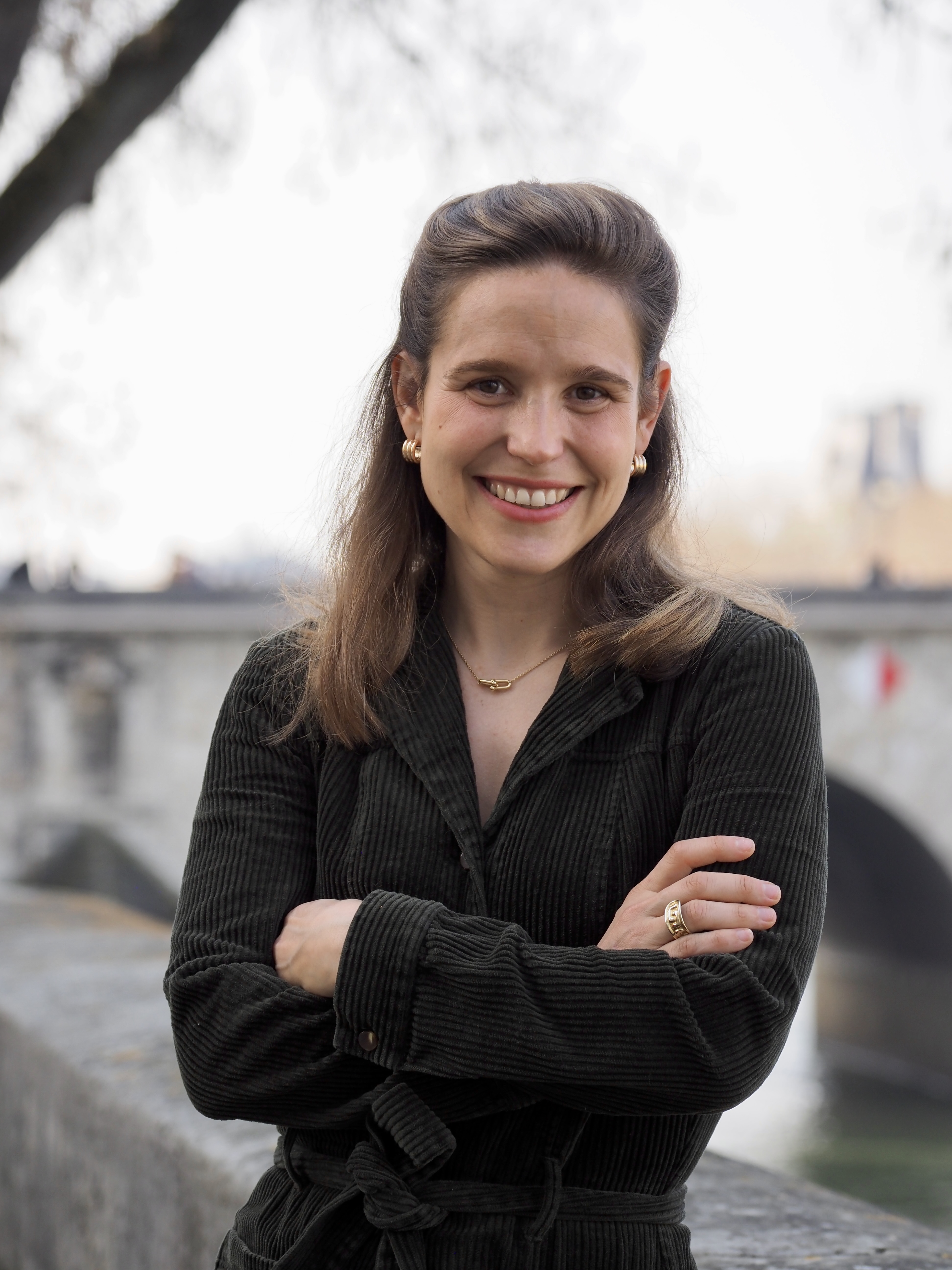 Femme souriante aux cheveux bruns portant un manteau noir, posant bras croisés près d'un pont.
