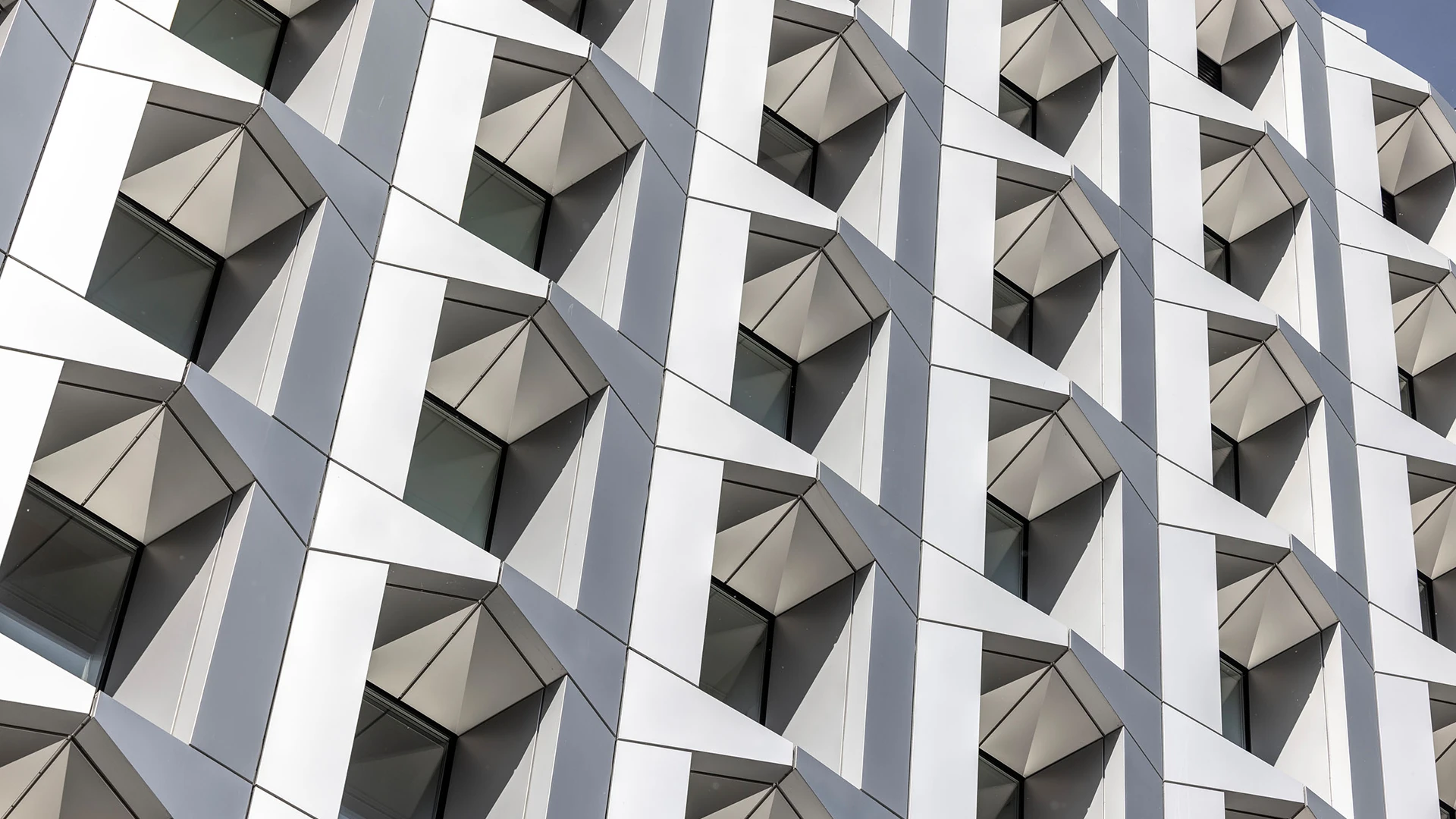 Modern building facade with geometric, angular white panels and dark windows arranged in a grid.
