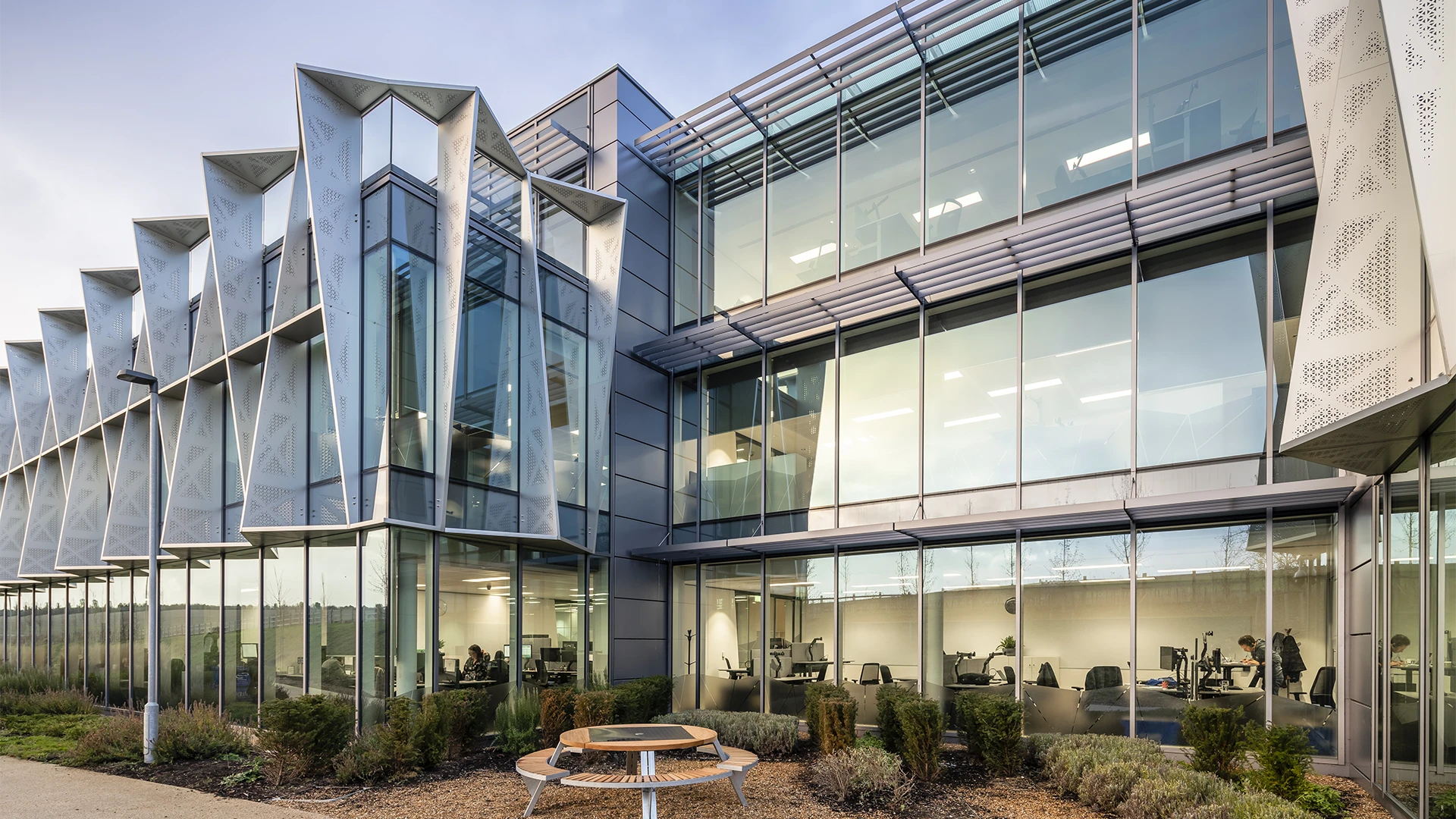 Modern glass office building with angular metal facade and outdoor wooden bench seating area.