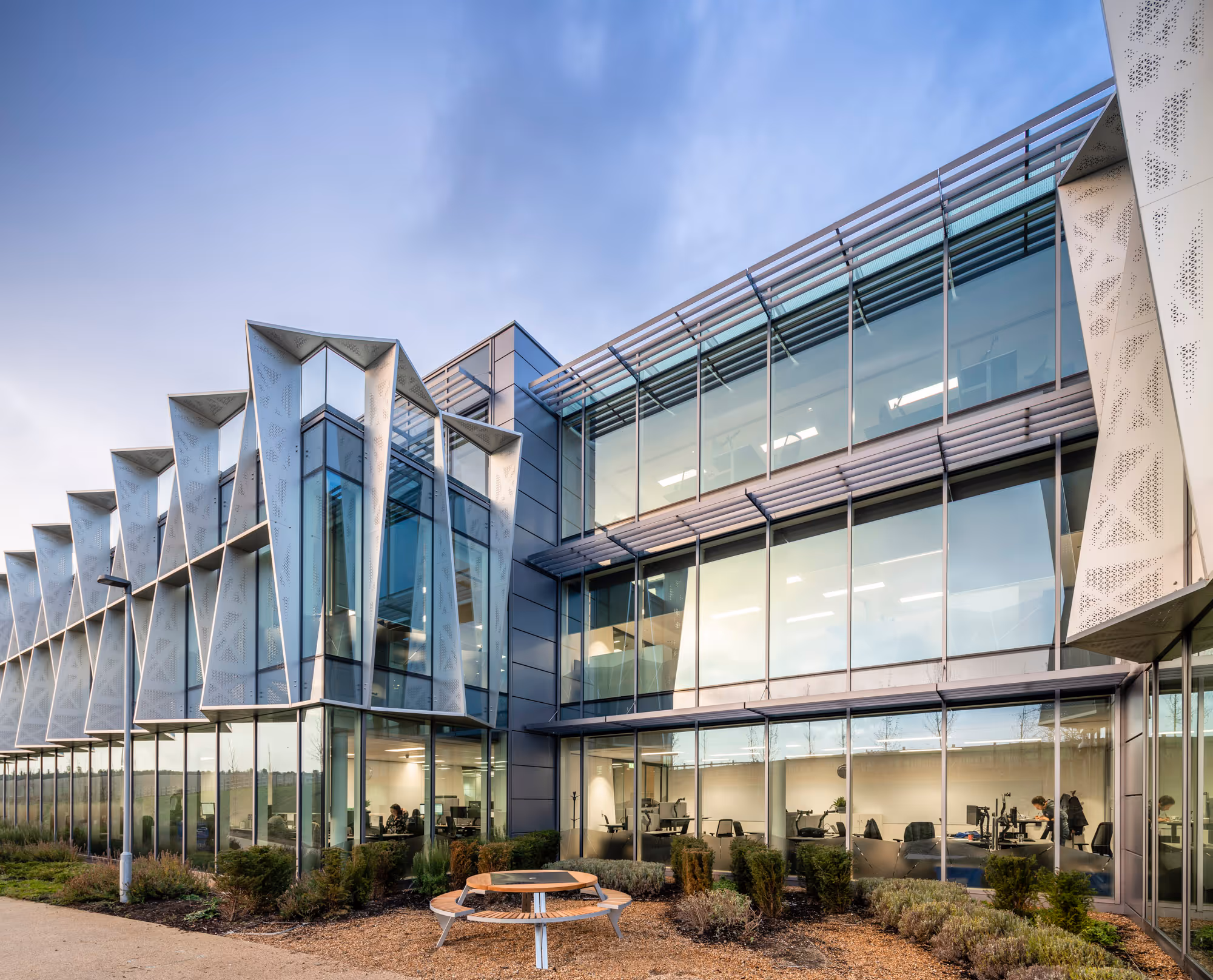 Modern glass office building with angular metal facade and outdoor wooden bench seating area.