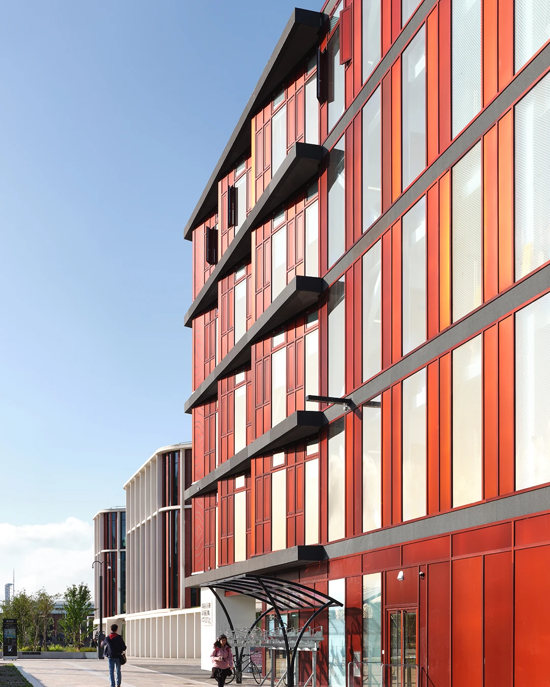 Modern red building with large windows and two pedestrians walking outside on a sunny day.