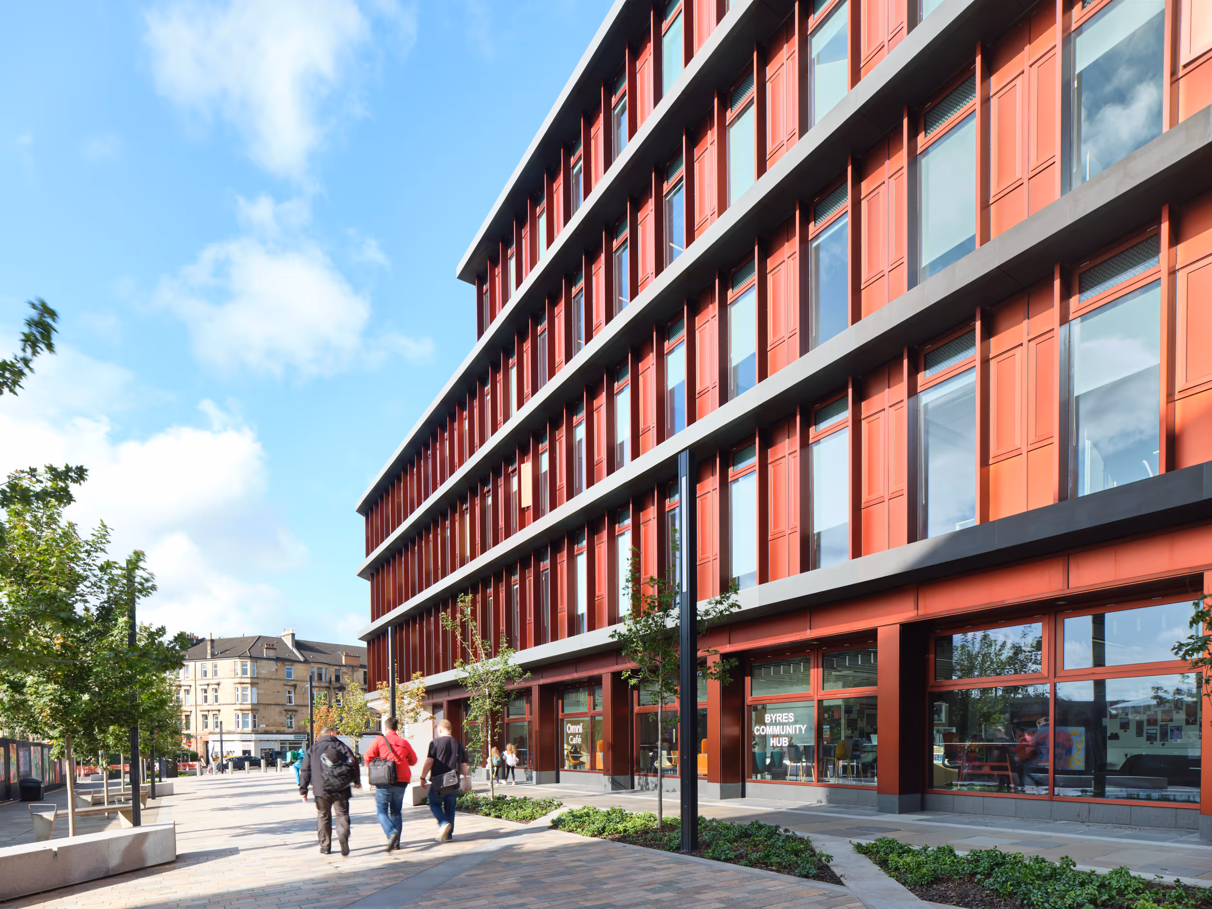 Modern red building with large windows beside a pedestrian walkway with three people walking by.