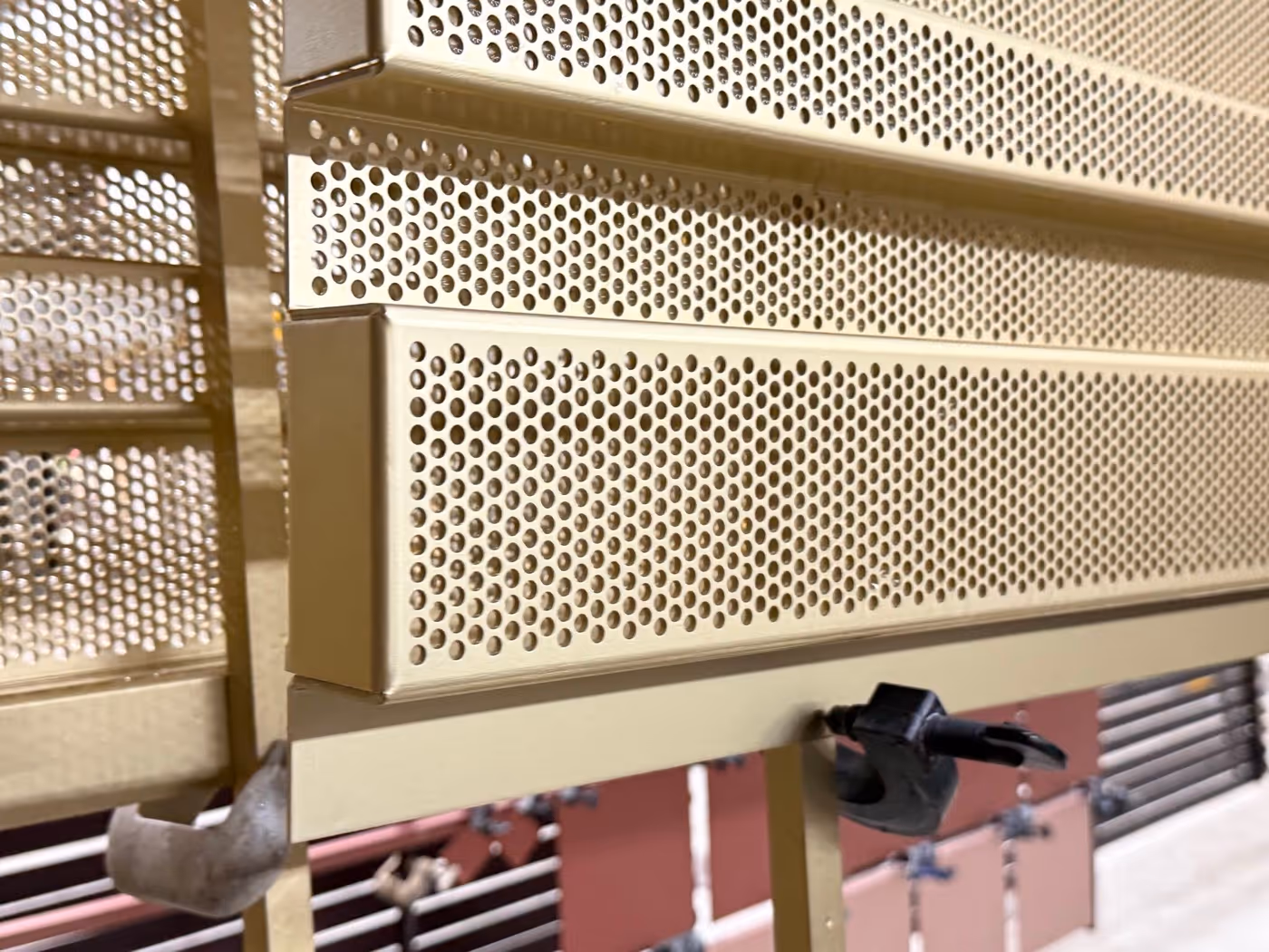 Close-up of beige perforated metal panels with a black clamp attached on a beige frame.