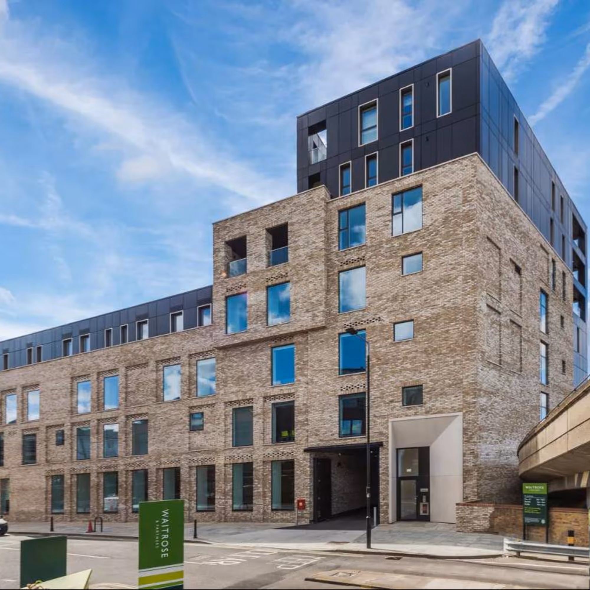 Modern multi-story brick and dark-paneled building under a blue sky, next to a Waitrose sign.