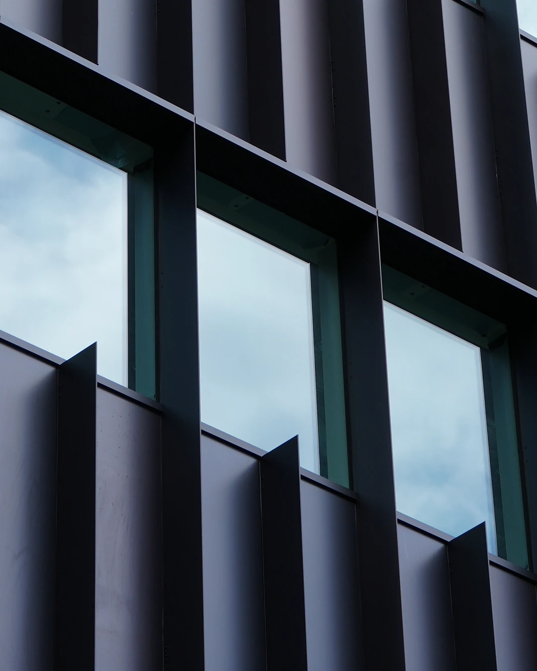 Close-up of a modern building facade with vertical black fins and rectangular windows reflecting sky.