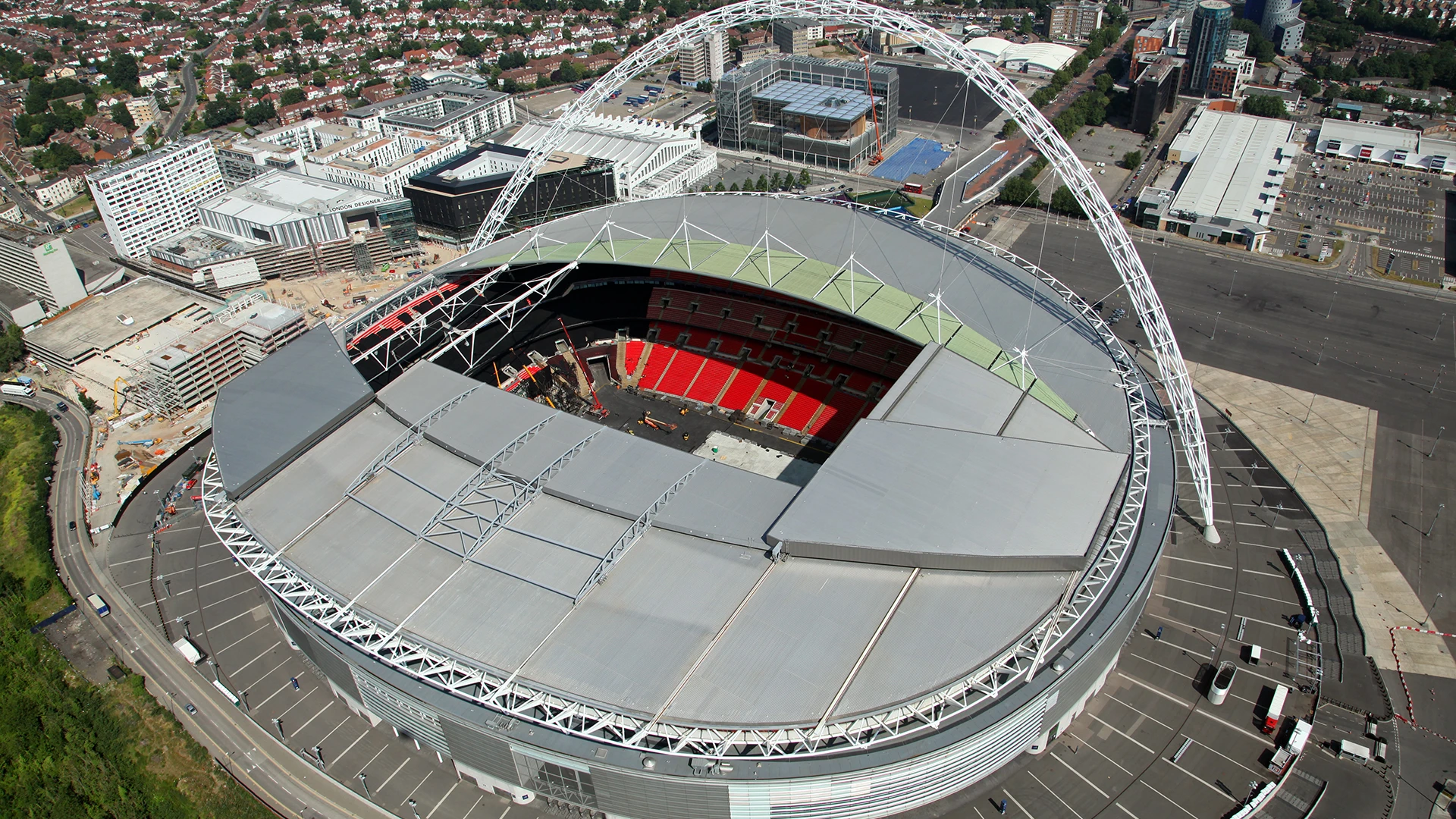 Aerial view of Wembley Stadium with its partial roof, red seats, and surrounding urban area.
