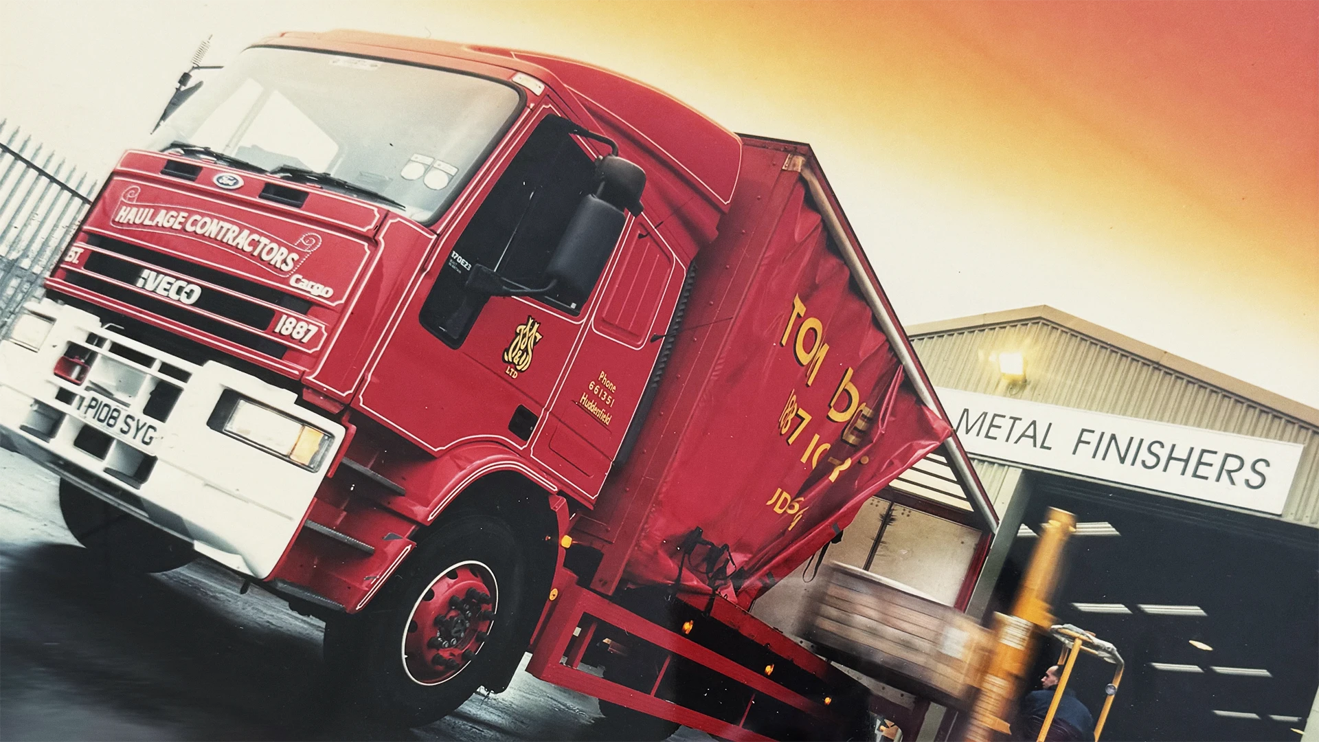 Red delivery truck beside warehouse with forklift unloading pallets near 'Metal Finishers' sign.