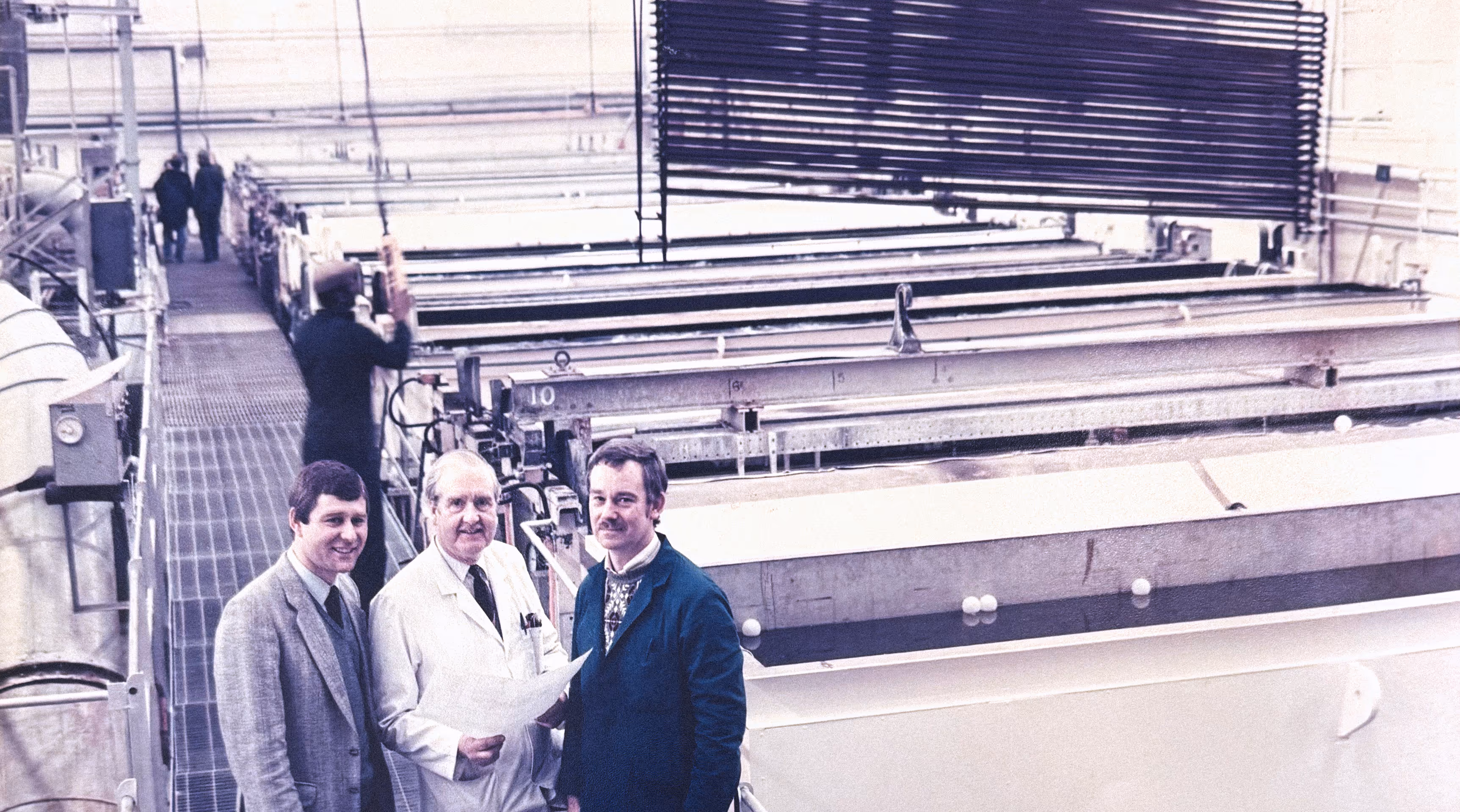 Three men standing in an industrial factory with metal equipment and conveyor belts.