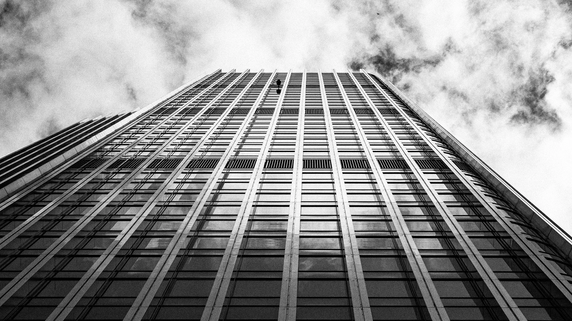 Black and white photo from below of a tall glass office skyscraper against a cloudy sky.