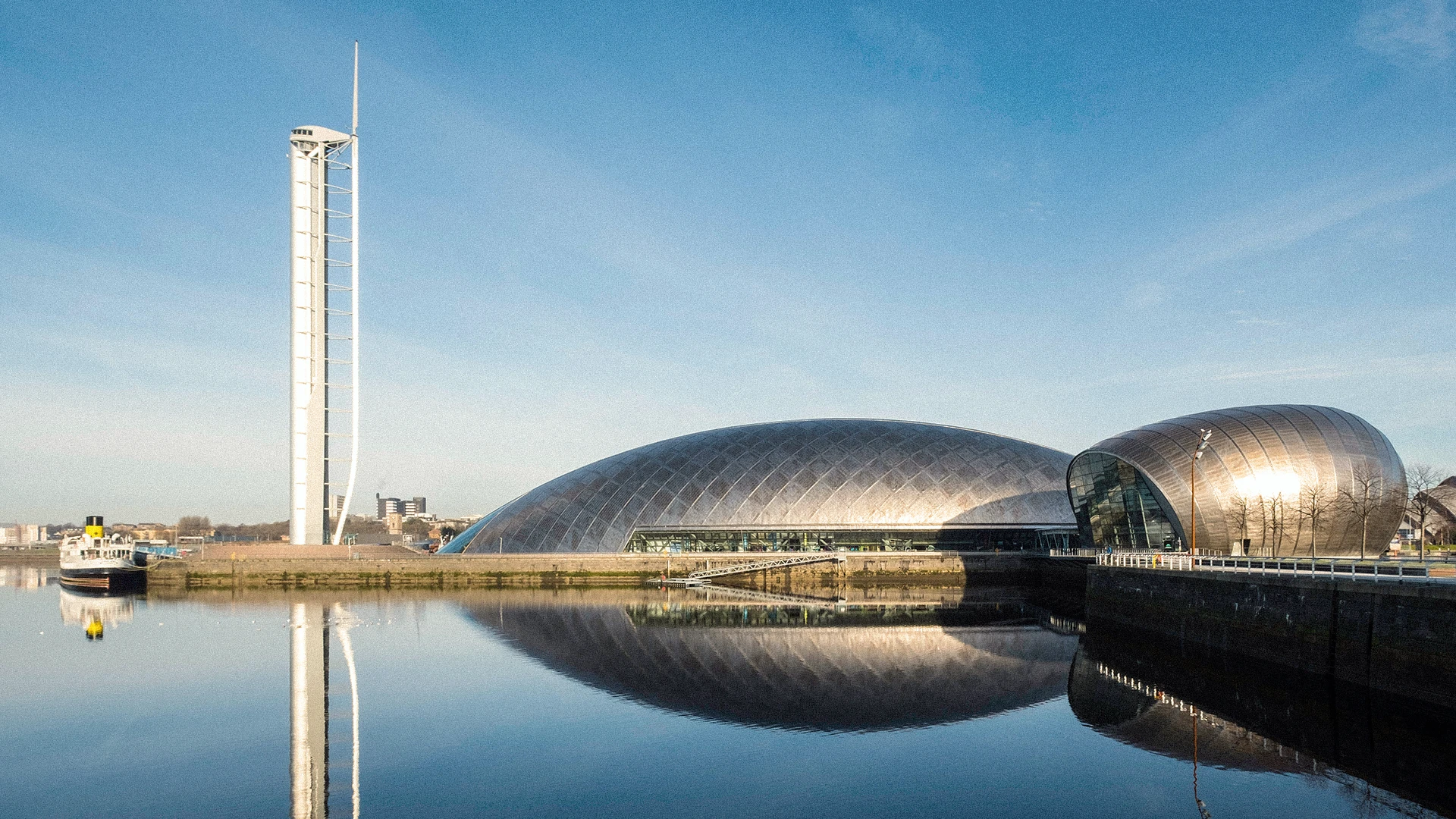 Modern curved buildings and a tall white tower reflected in calm water under clear blue sky.