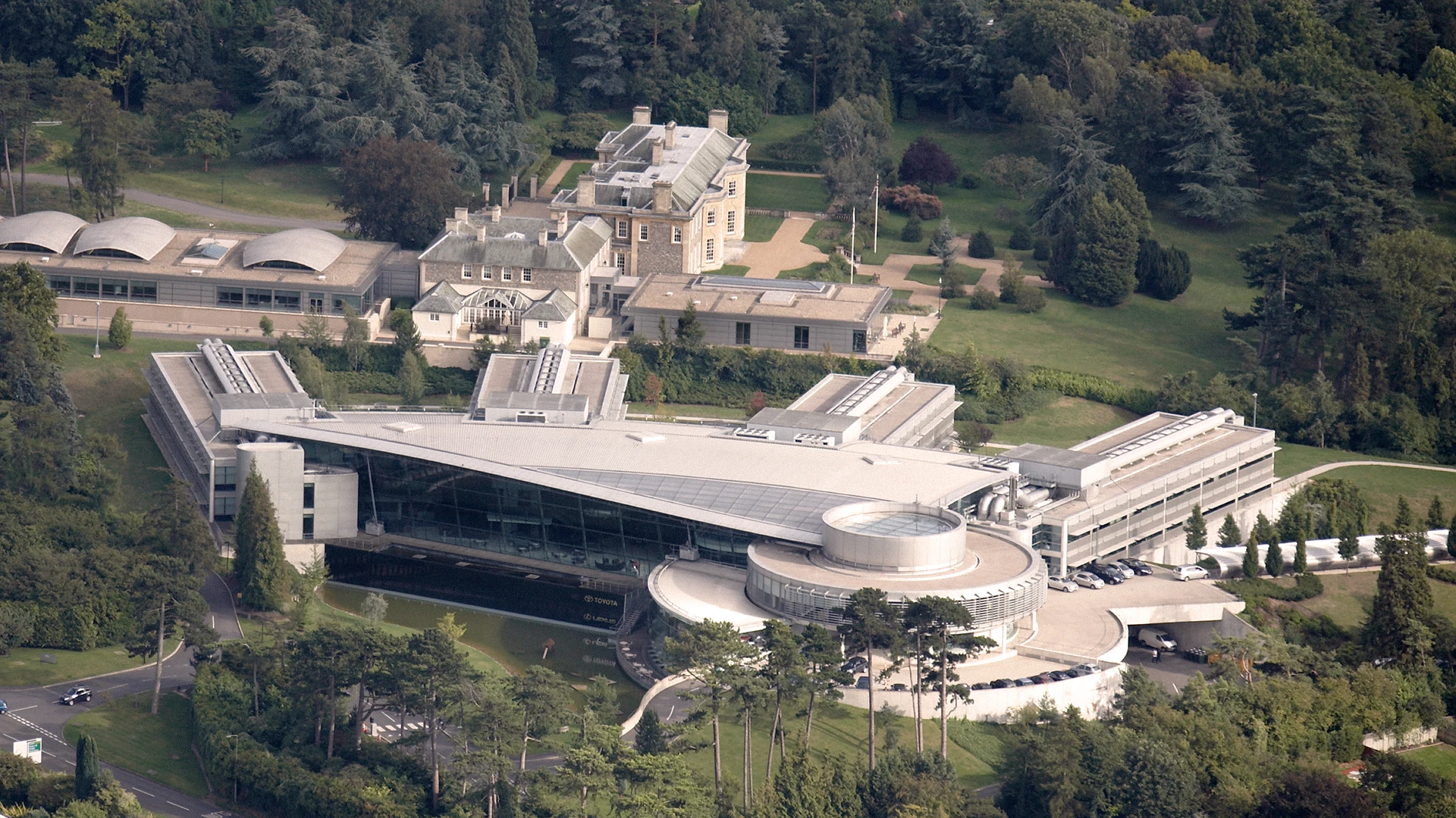 Aerial view of a large modern building with circular and angular sections surrounded by trees and lawns.