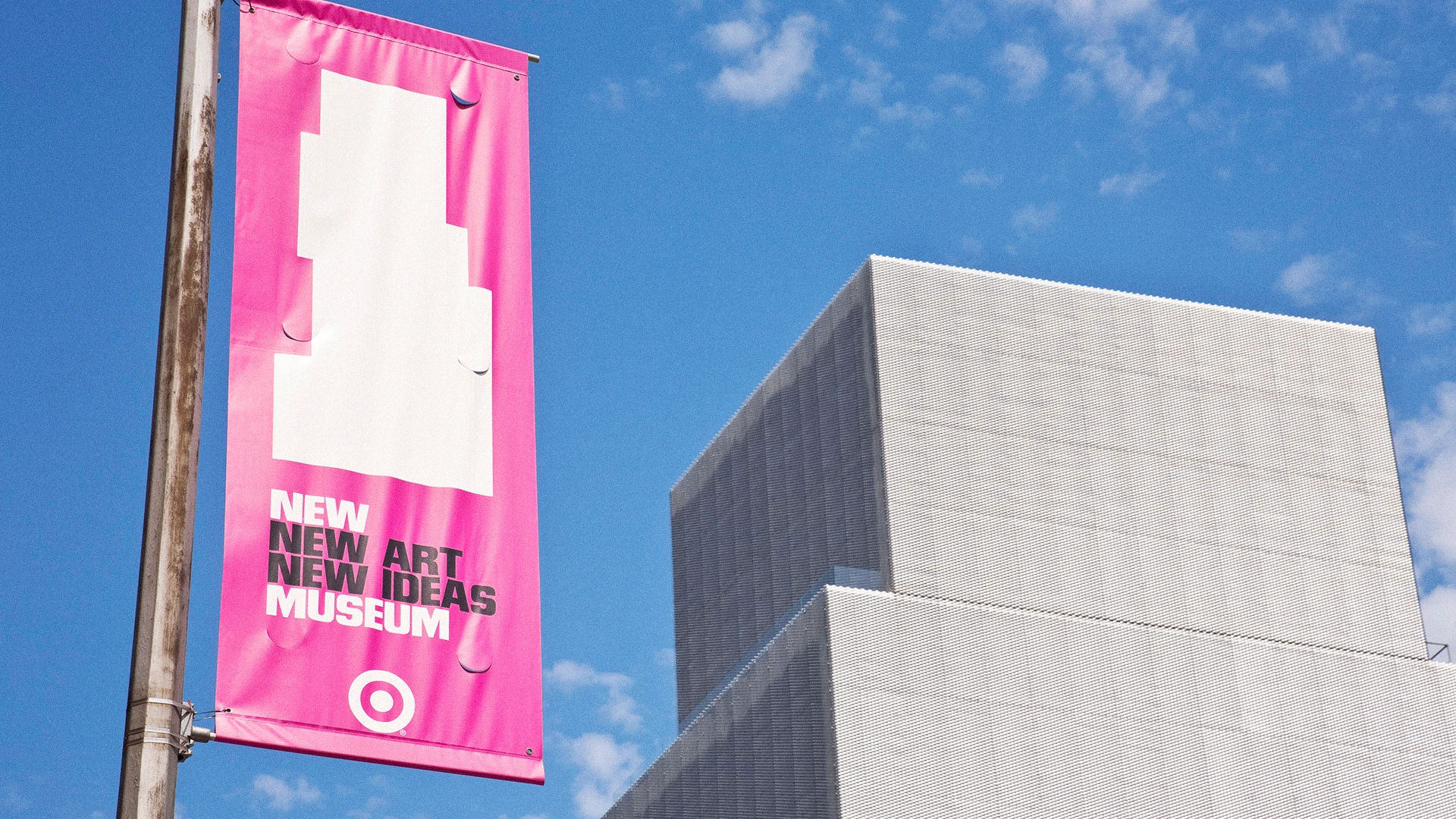 Pink banner on pole reads New Art New Ideas Museum against a blue sky and modern building.