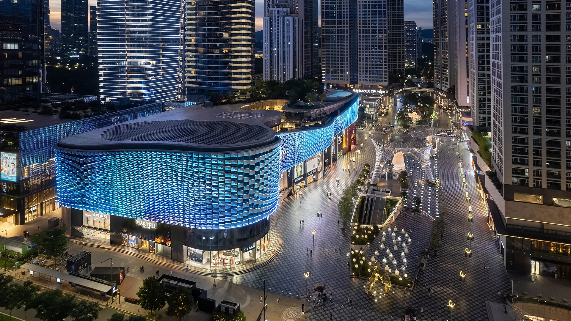 Night view of a modern shopping plaza with blue-lit facade and pedestrian plaza with art installations.
