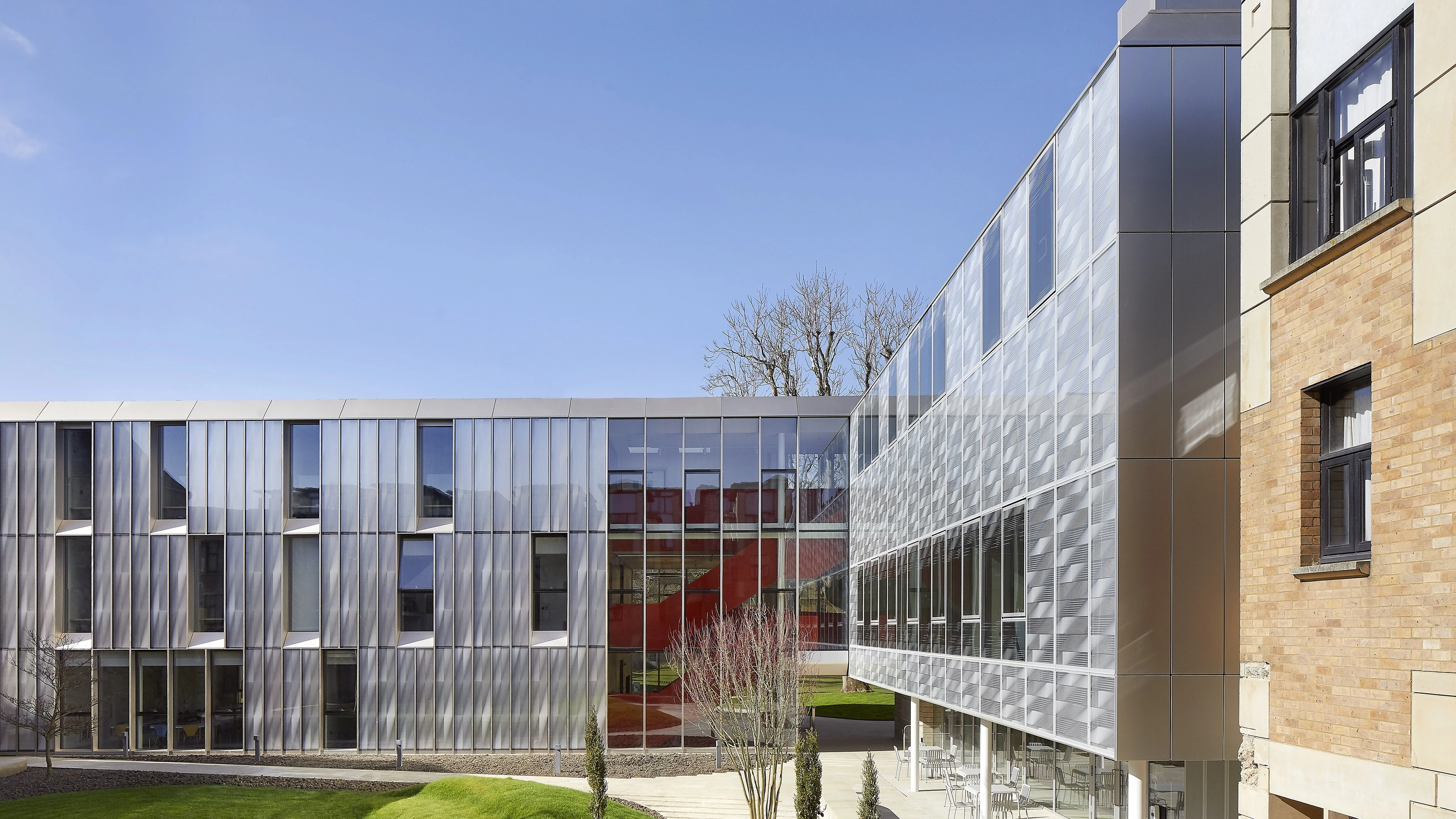 Modern building with glass and metal facade, red interior staircase, and landscaped grassy courtyard.