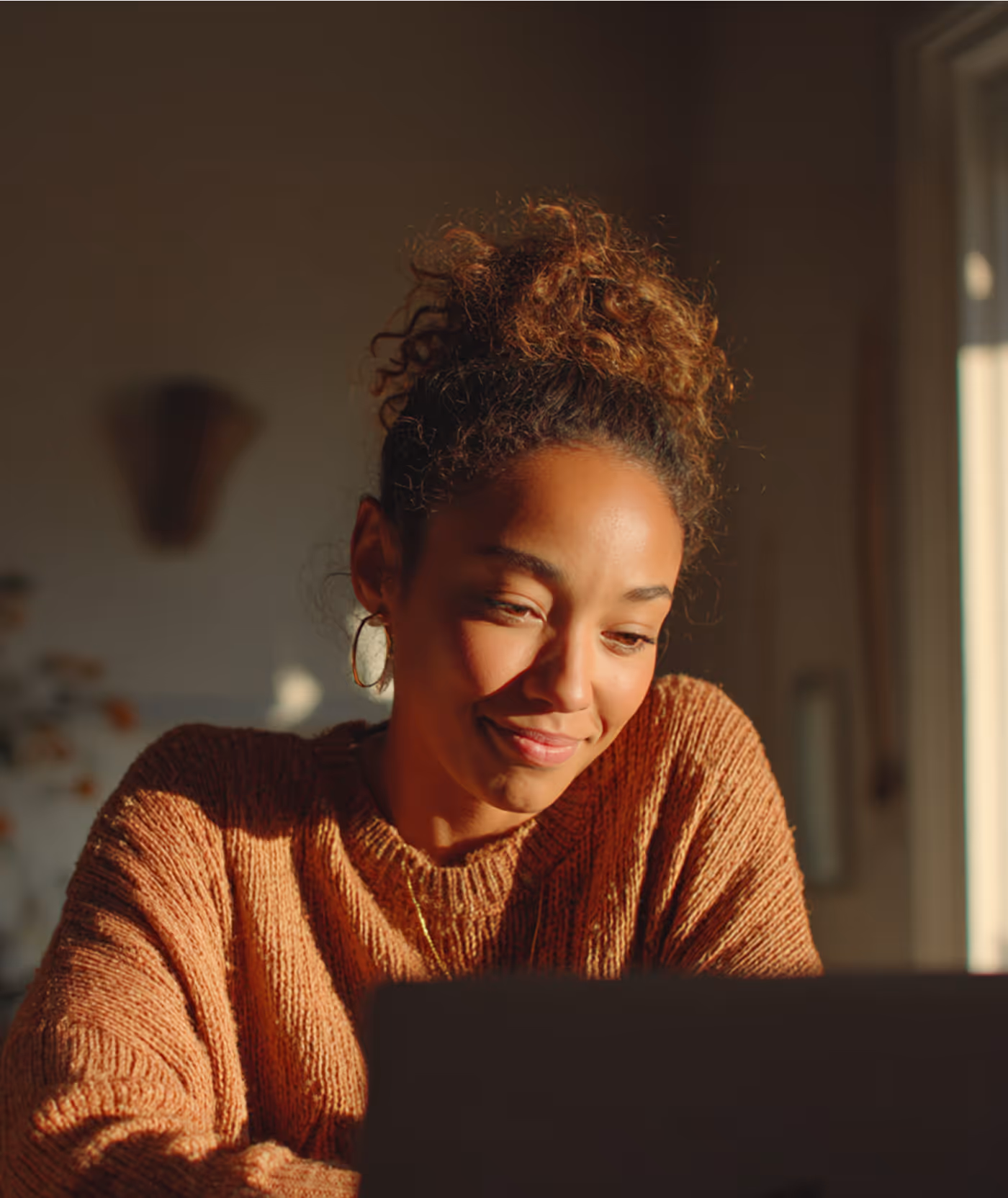 Jeune femme souriante portant un pull en maille marron, assise devant un ordinateur dans une pièce lumineuse.