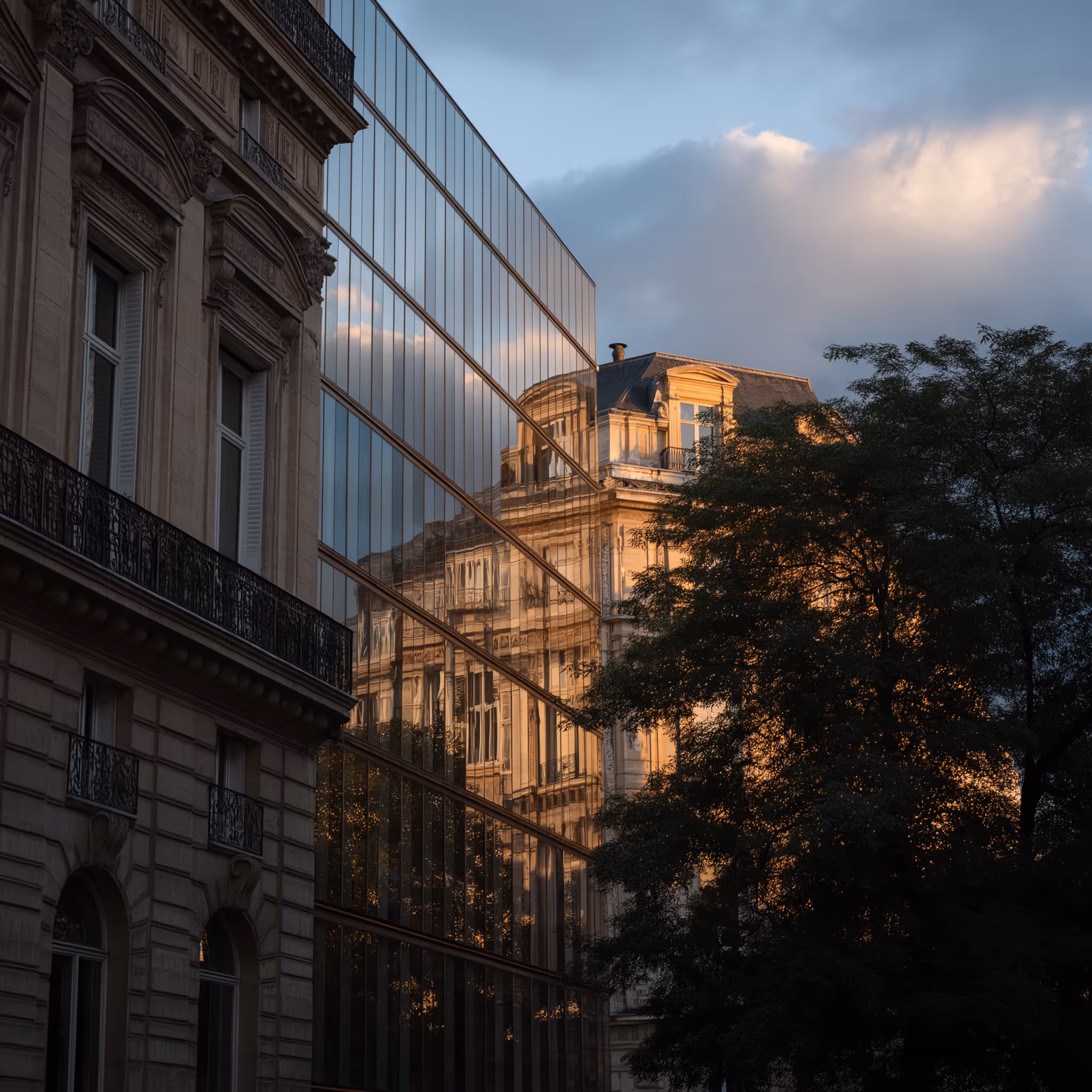 Façade d'un bâtiment ancien reflétée sur les vitres d'un immeuble moderne à côté d'un arbre, au coucher du soleil.