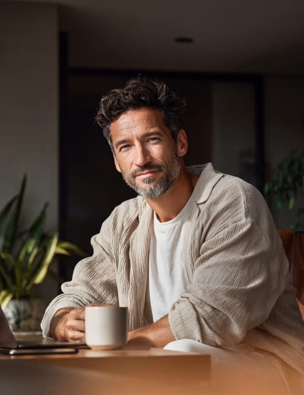 Homme souriant avec barbe grisonnante assis à une table avec une tasse dans un intérieur moderne.
