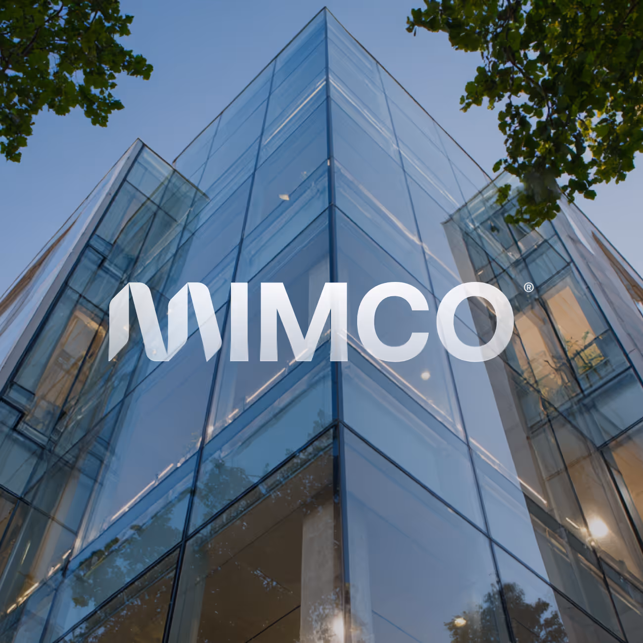 Façade en verre d'un bâtiment moderne avec le logo MIMCO au centre, entouré d'arbres verts.