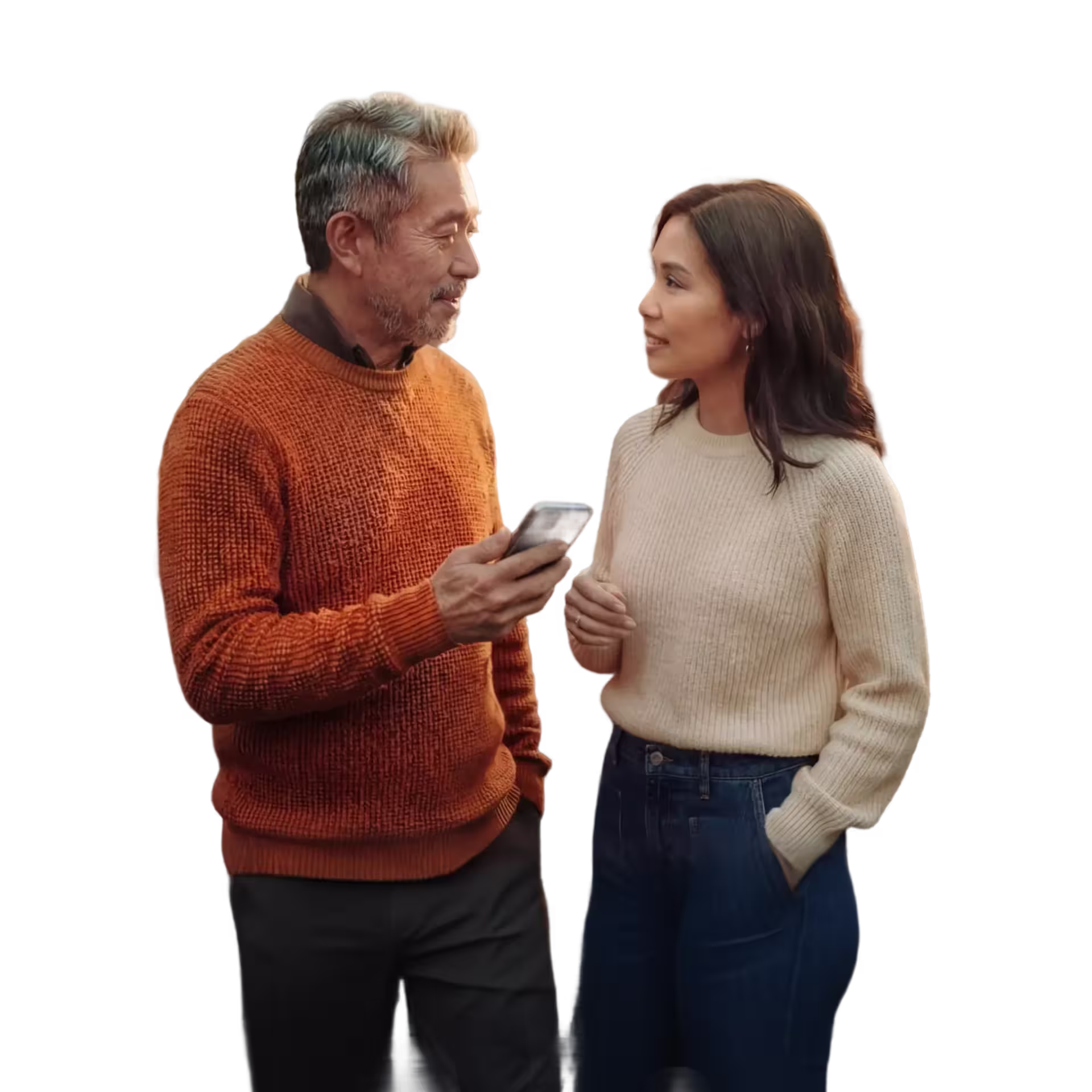 Middle-aged man in orange sweater shows a smartphone to a woman in a cream sweater and jeans as they engage in conversation.